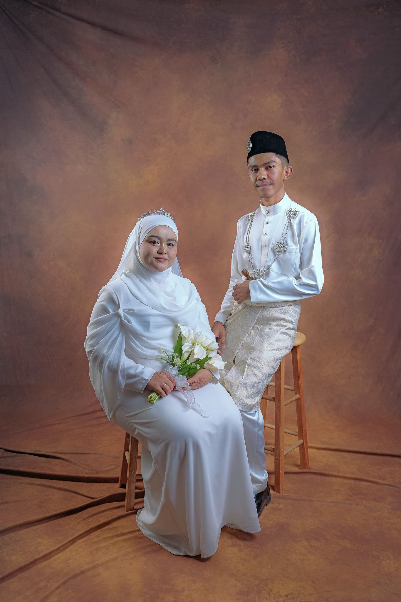 Traditional Malay wedding portrait showing couple in white traditional attire sitting and standing on wooden stools.