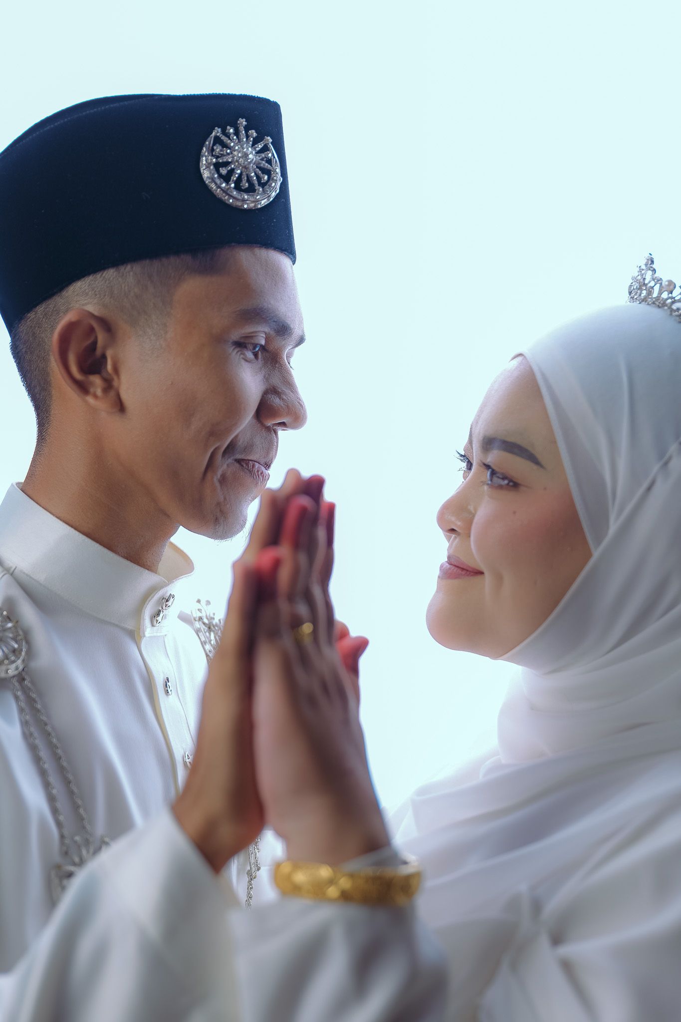 A couple in traditional Muslim wedding attire exchange sweet glances in a bright white setting during their special day.