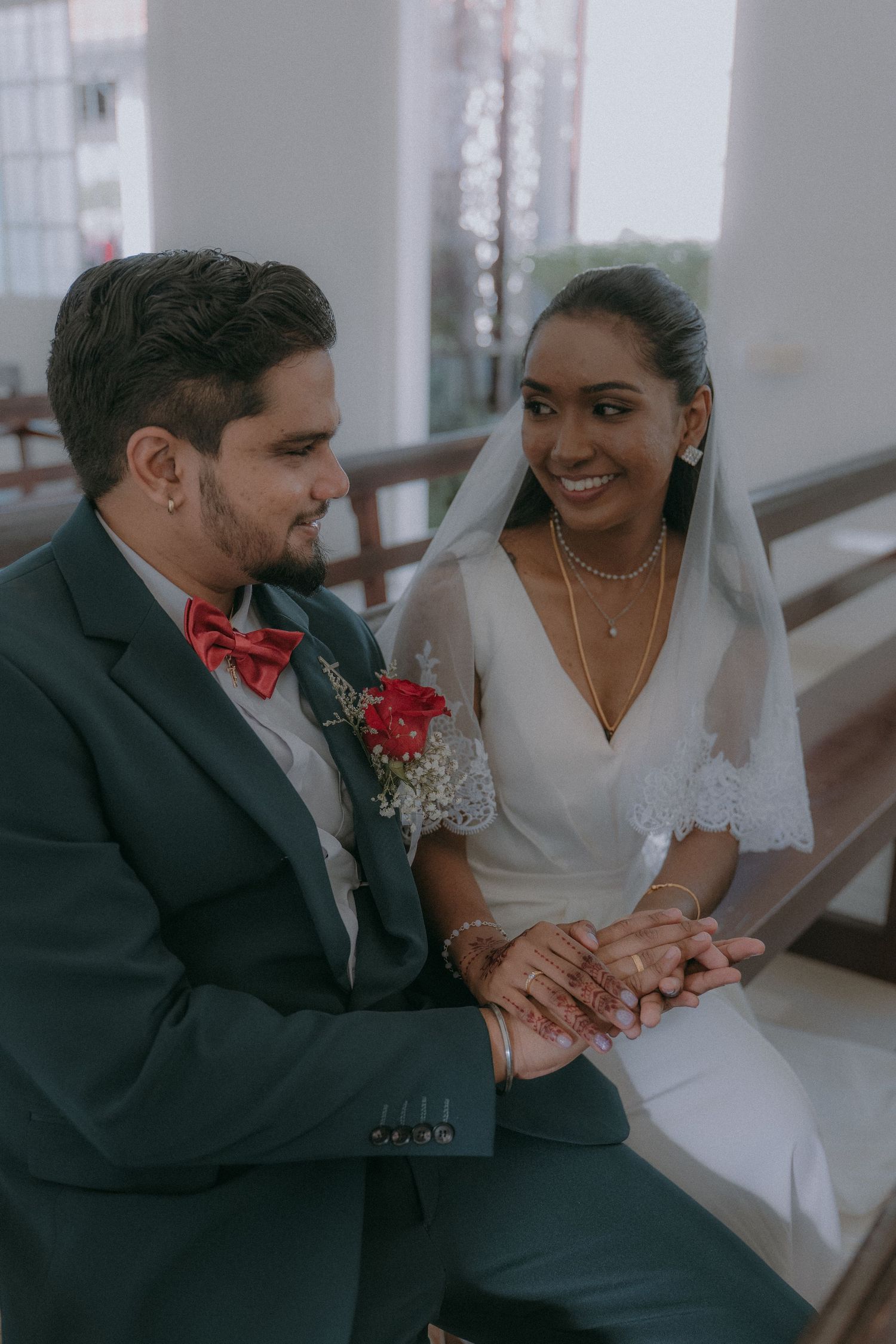 Newlywed couple share intimate moments seated together on bench while holding hands and smiling.