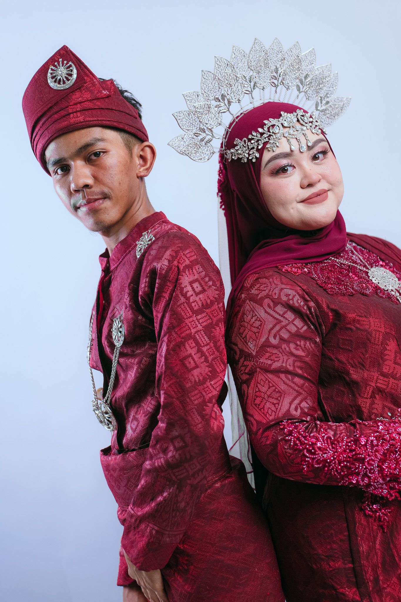 Traditional Malay wedding couple wearing matching red songket outfits and elaborate accessories stand back-to-back.