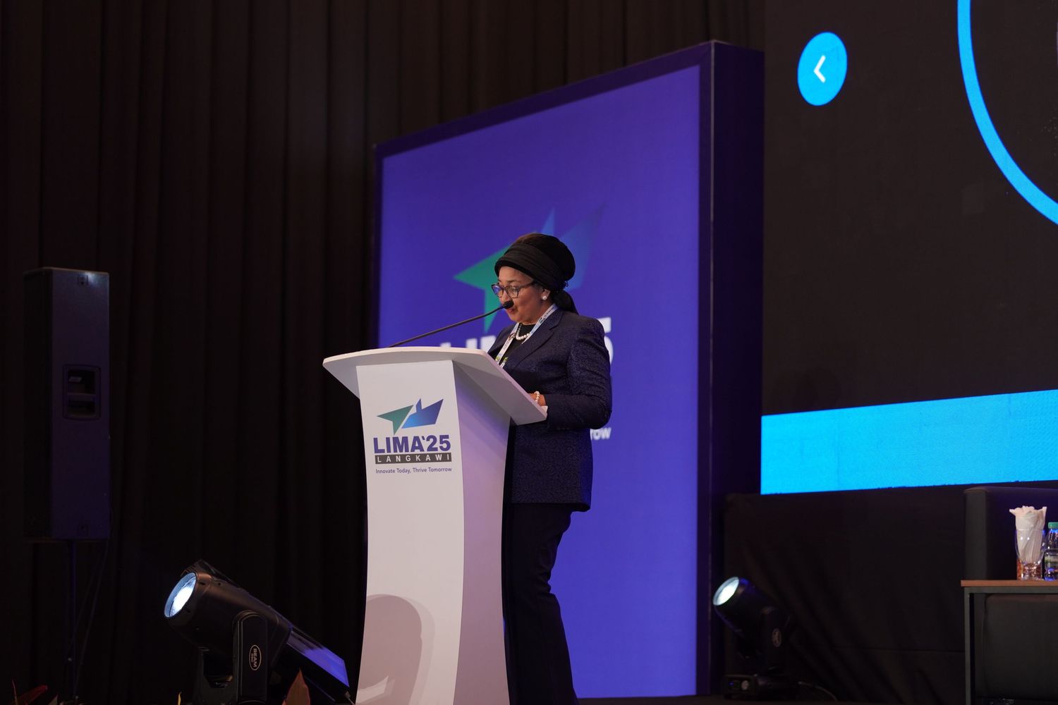 Professional delivering speech at white podium with blue digital display in background.