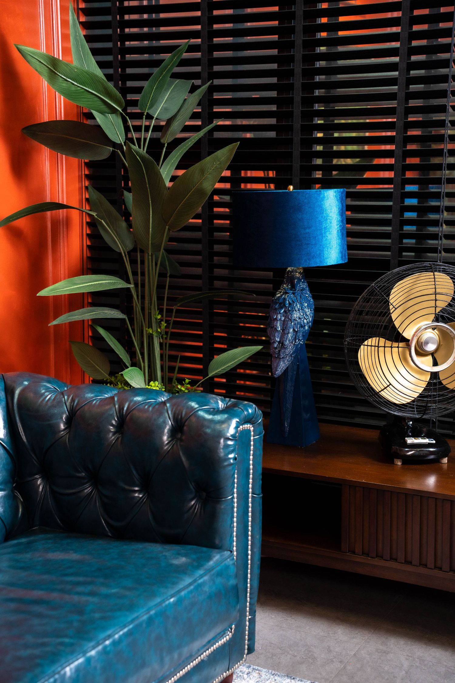 Modern blue leather couch with studded details sits against dark blinds with ambient lighting and tropical plants.
