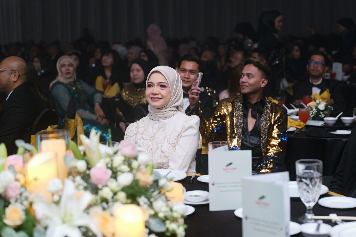 Elegant woman in white hijab seated at gala dinner table with floral centerpiece arrangement.