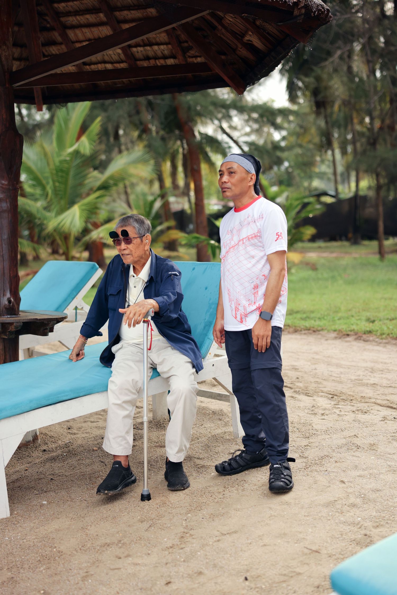 Elderly Asian man with cane sitting on beach lounger assisted by caregiver at tropical resort.