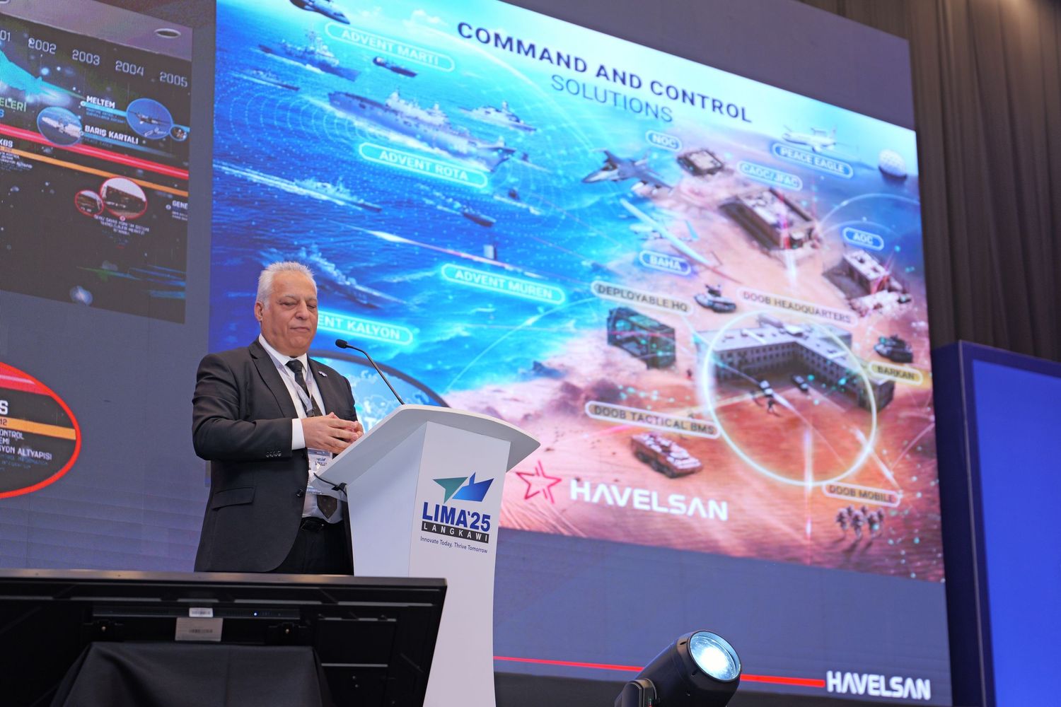 Speaker presents at podium with maritime defense strategy slide displayed on screen.