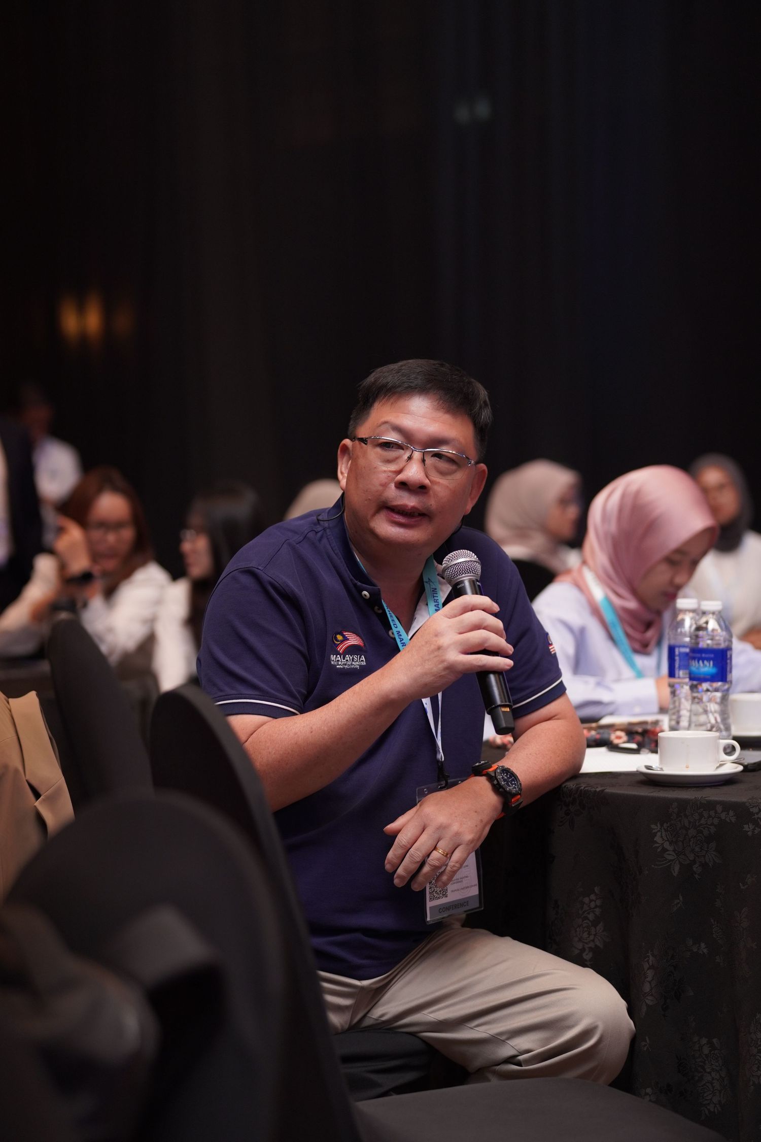 Speaker in navy polo shirt addressing audience at round table conference setting.