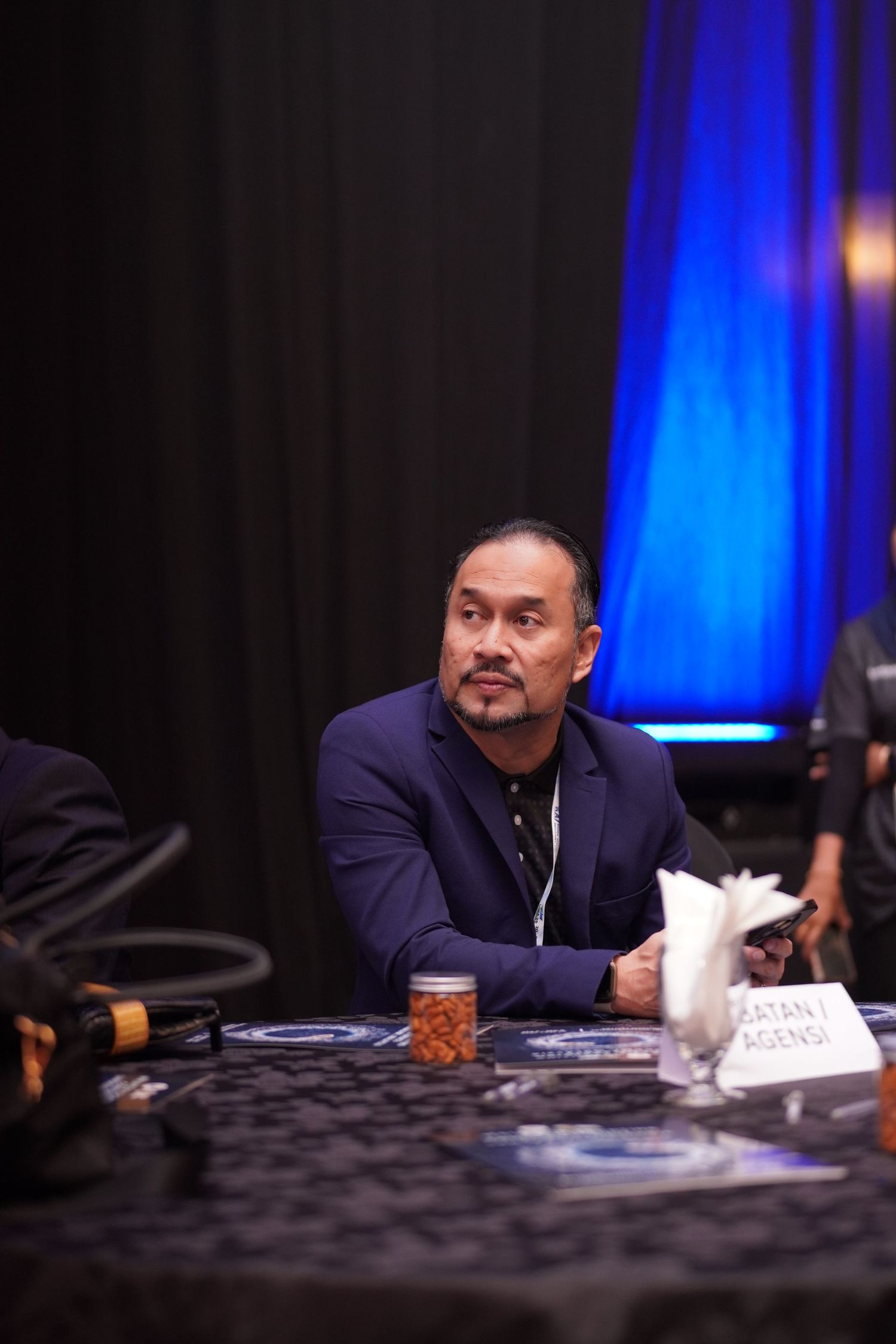 Conference attendee in a dark suit sits at a table with blue lighting in the background during a business event.