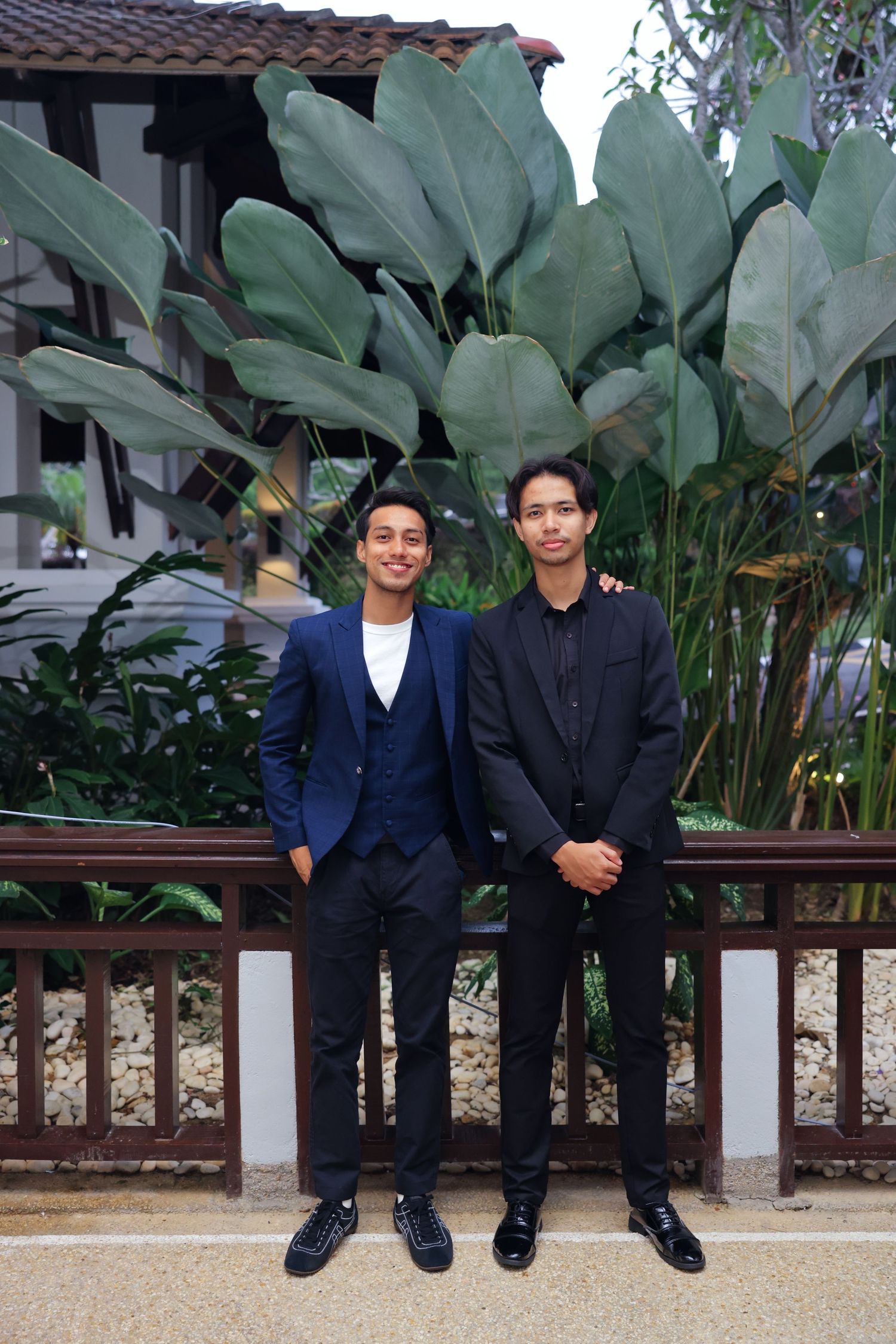 Two young men in formal suits pose outdoors in front of large tropical plants and a wooden fence.