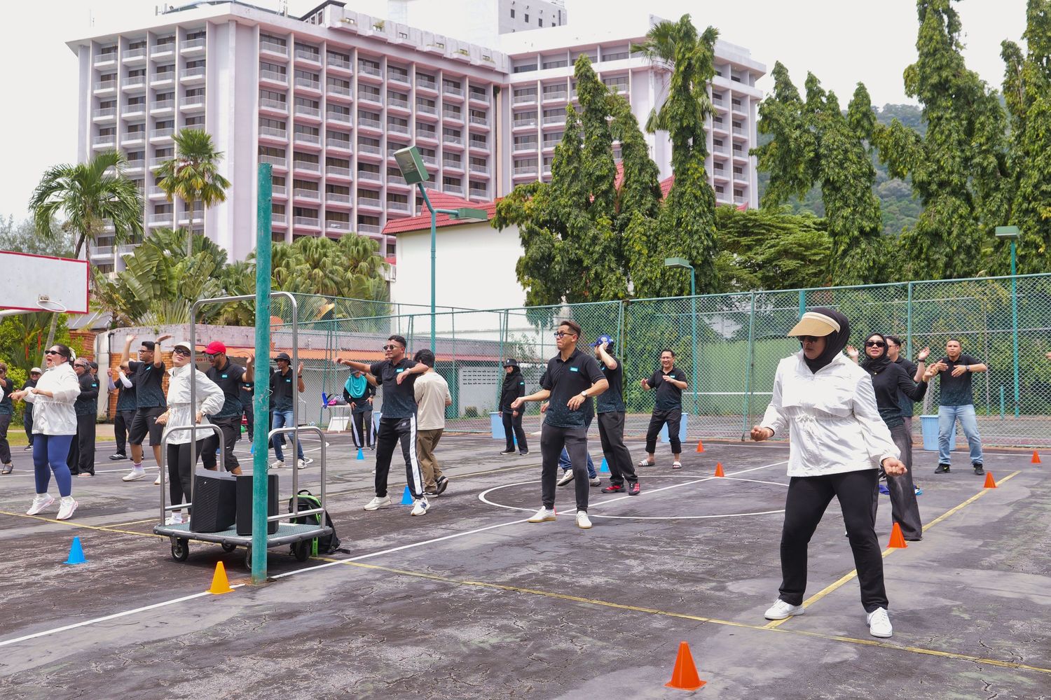 Group of people in black uniforms exercising outdoors on a sports court with orange cones and a large building backdrop.