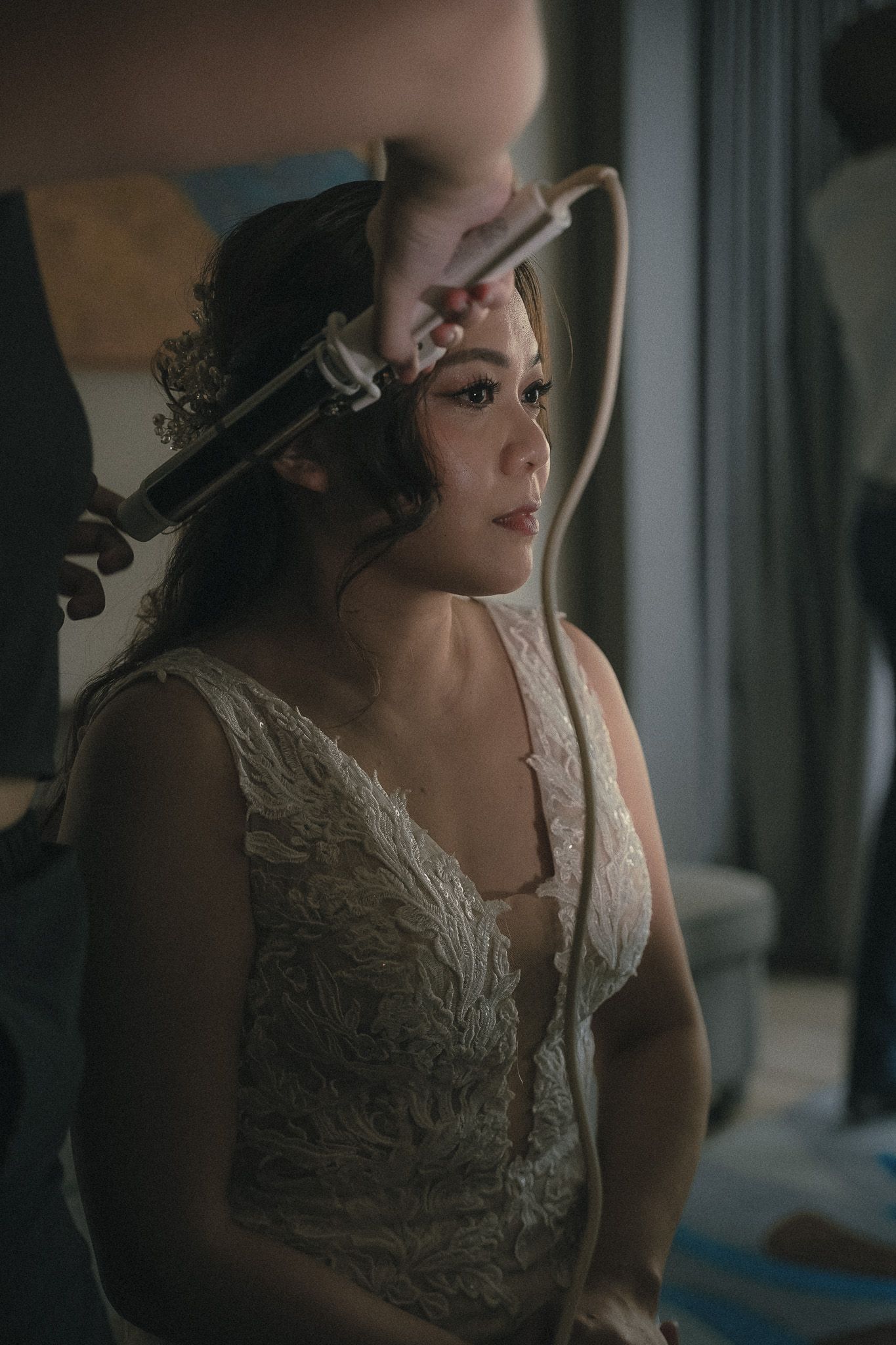 A bride in an elegant lace wedding dress getting ready by the window during morning preparations.