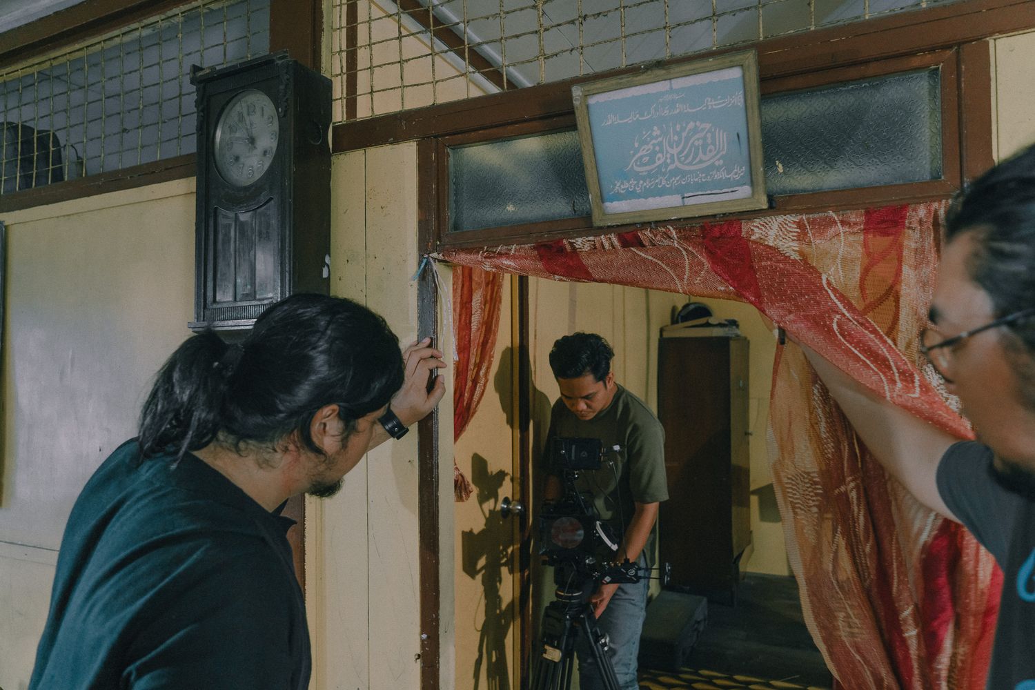 Film crew members reviewing footage together in a dimly lit room with vintage decor and a wall clock.