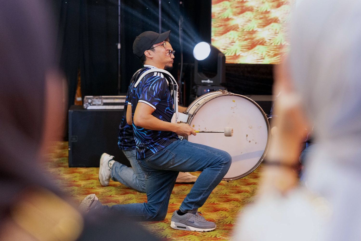Young man playing a large bass drum on one knee at an indoor music event or concert performance.
