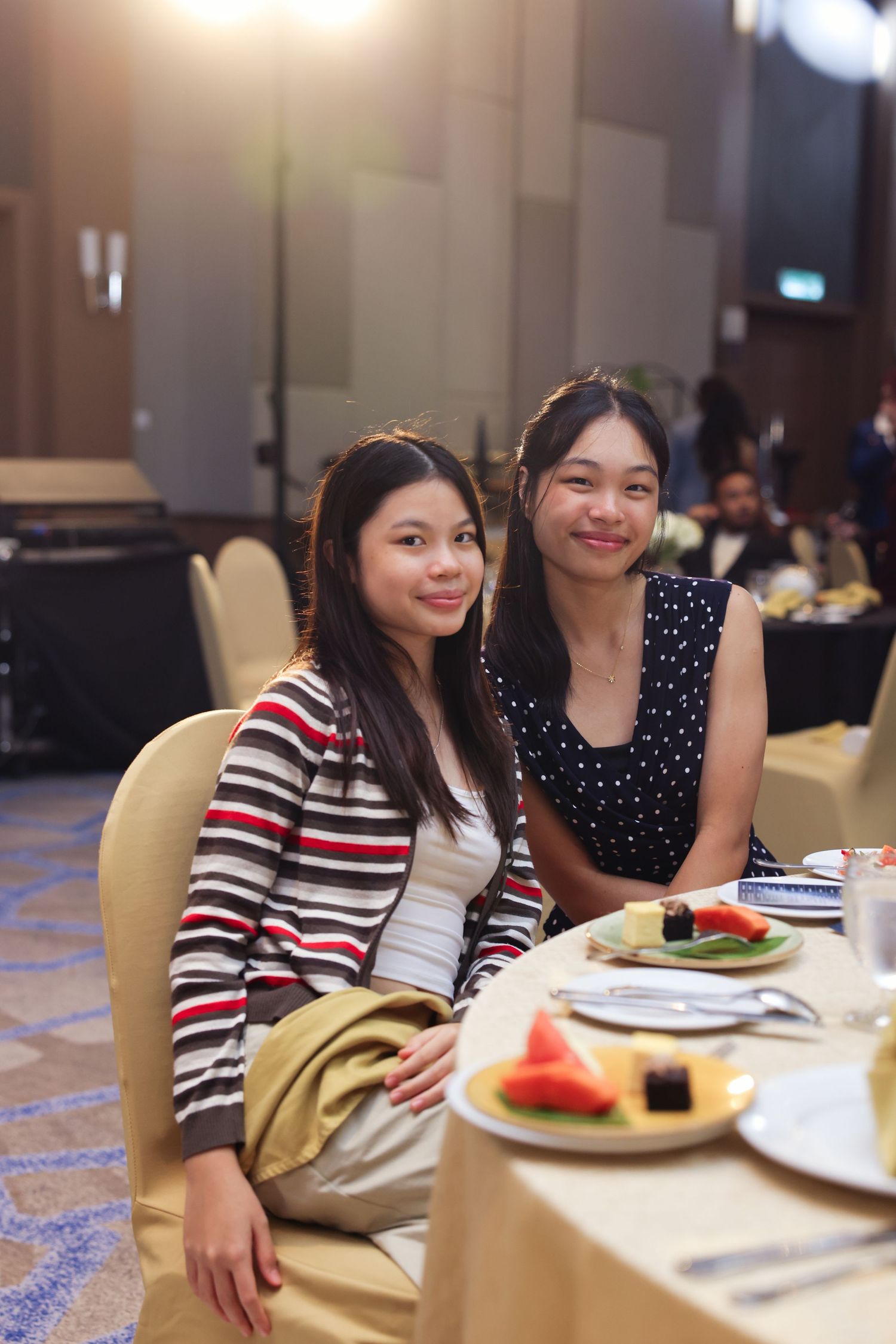 Two young women smile at a banquet table with fruit plates at a formal corporate dinner.
