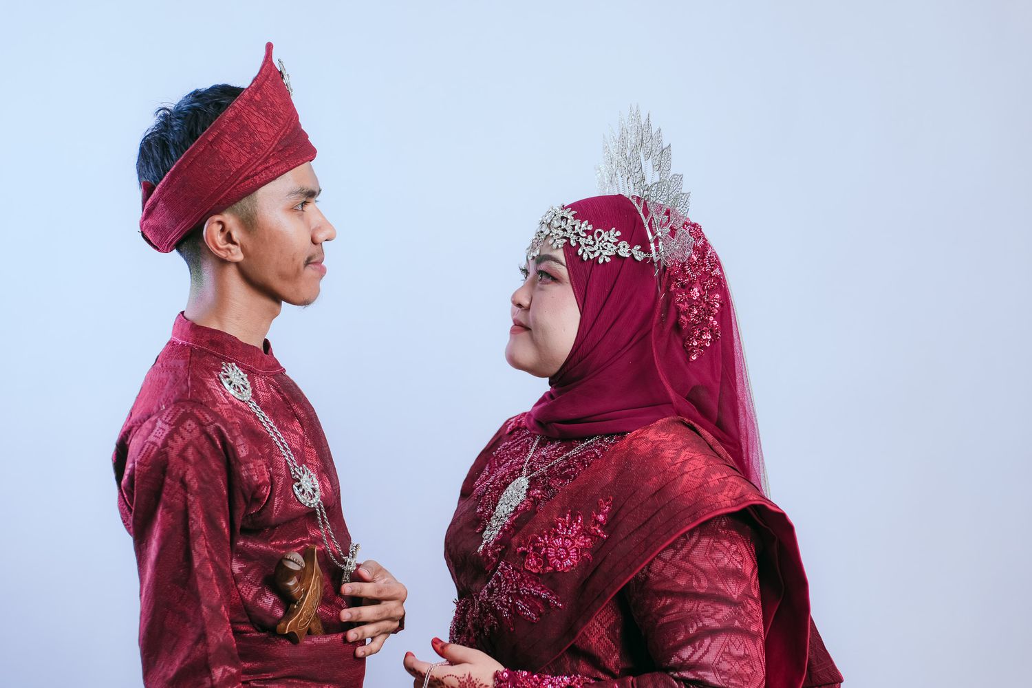 A couple in traditional red Malay wedding attire stand together against a white background.