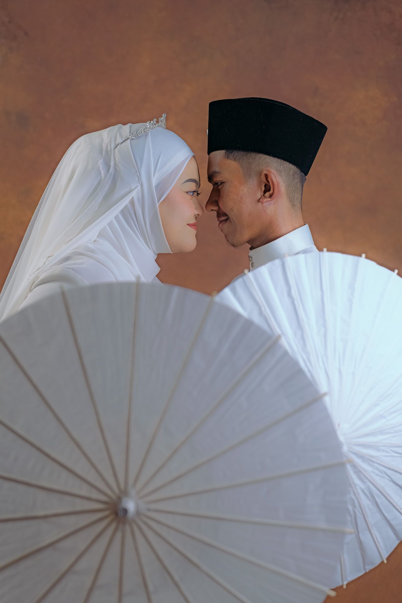 Traditional Malay wedding couple pose intimately behind a white parasol in a romantic portrait session.