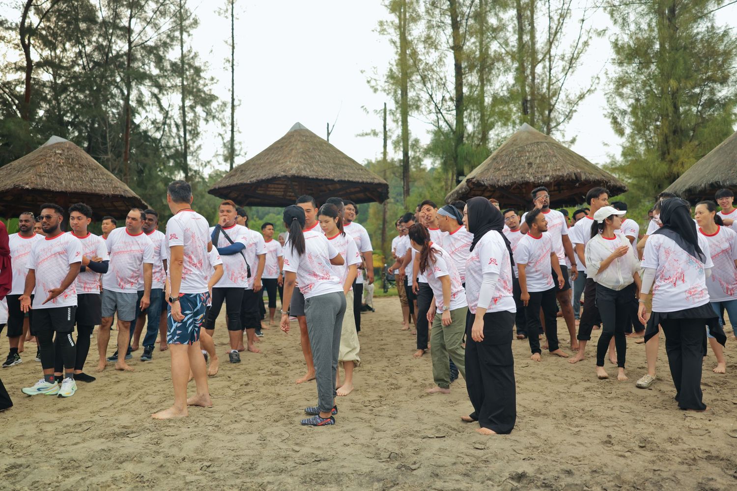 Large group of people in white t-shirts gathered on sandy beach near thatched huts and pine trees at outdoor event.
