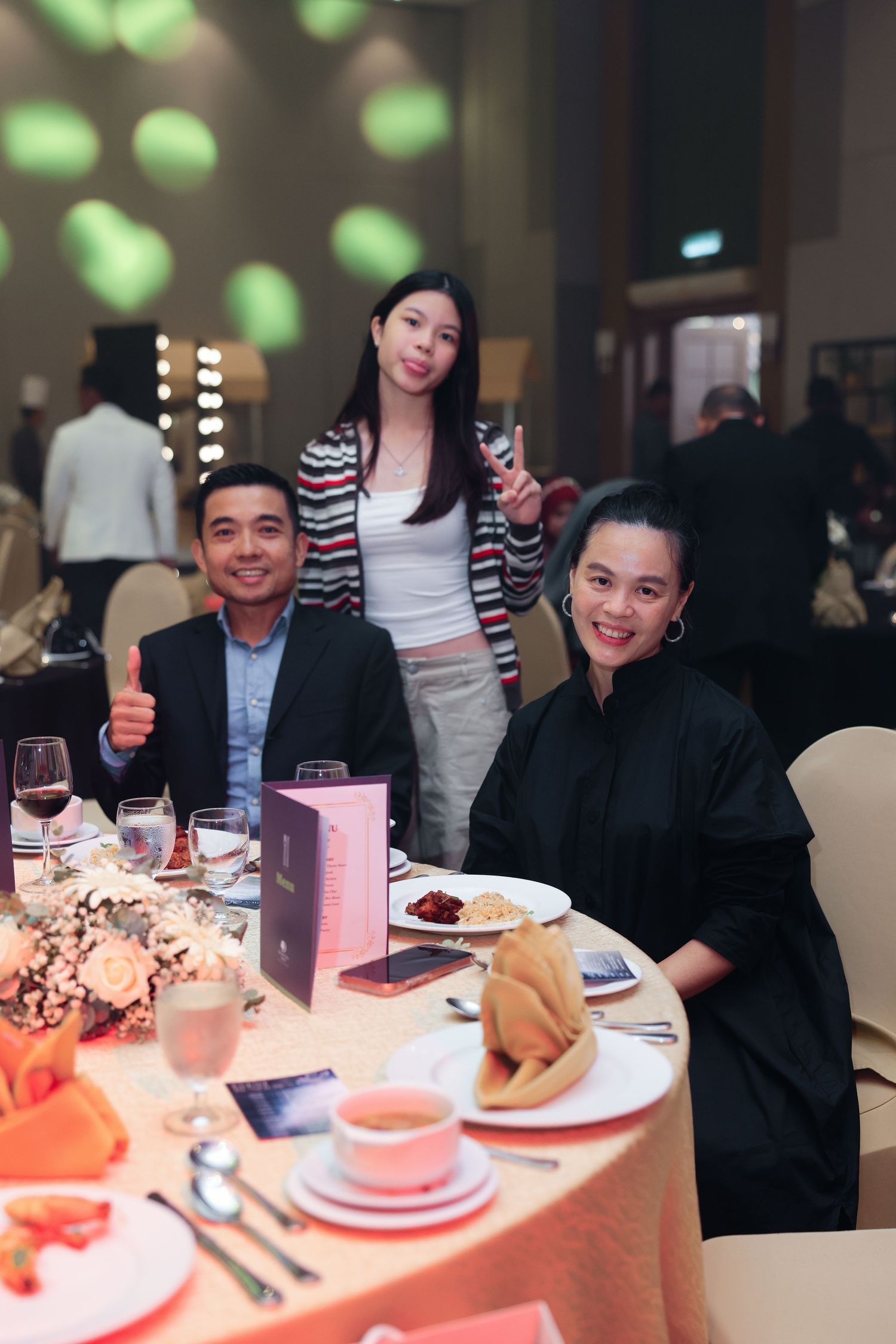 Three people pose at an elegant banquet table with floral centerpiece, plates, and glassware in a formal dining hall.