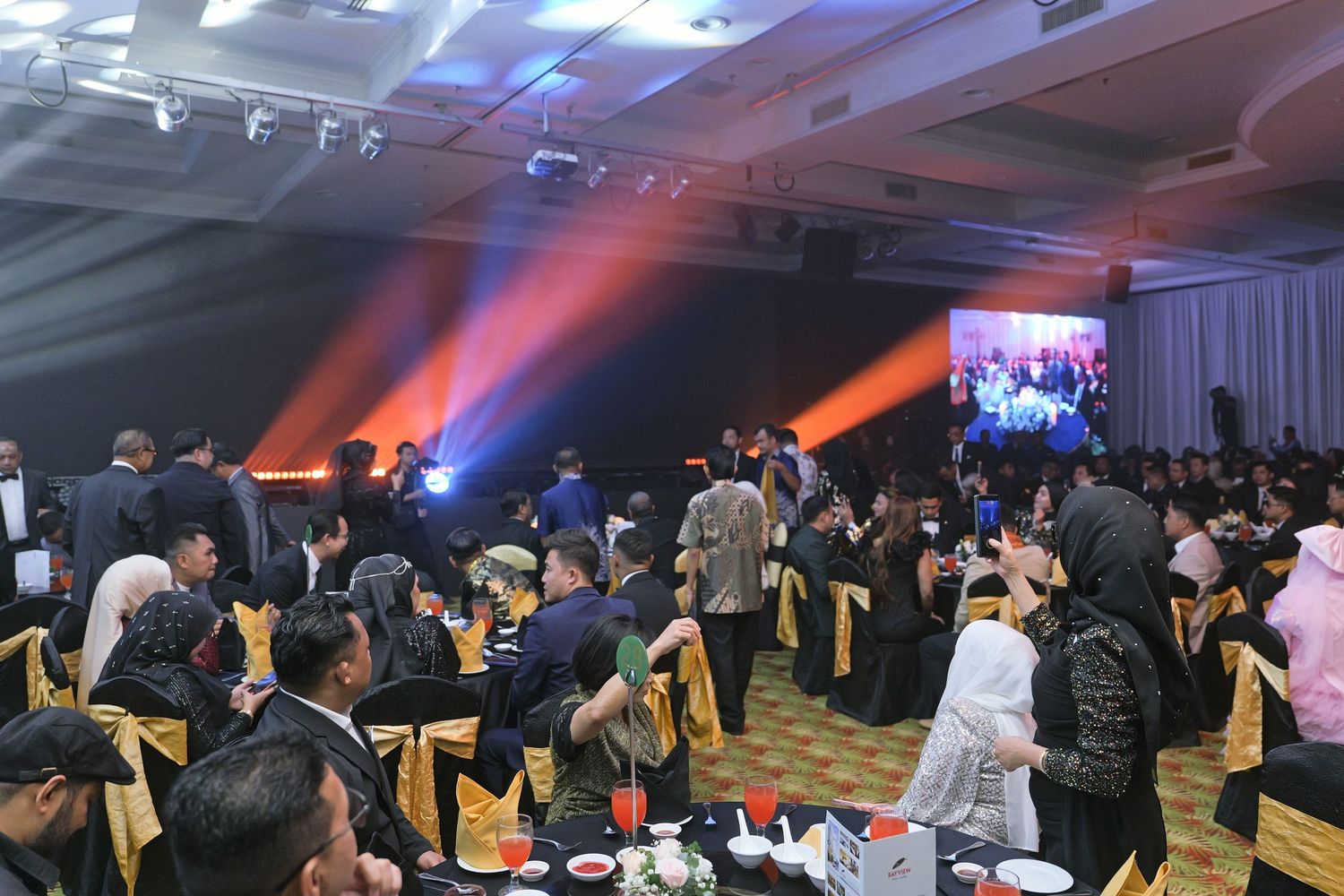 Large corporate gala dinner with guests seated at tables under dramatic orange stage lighting.