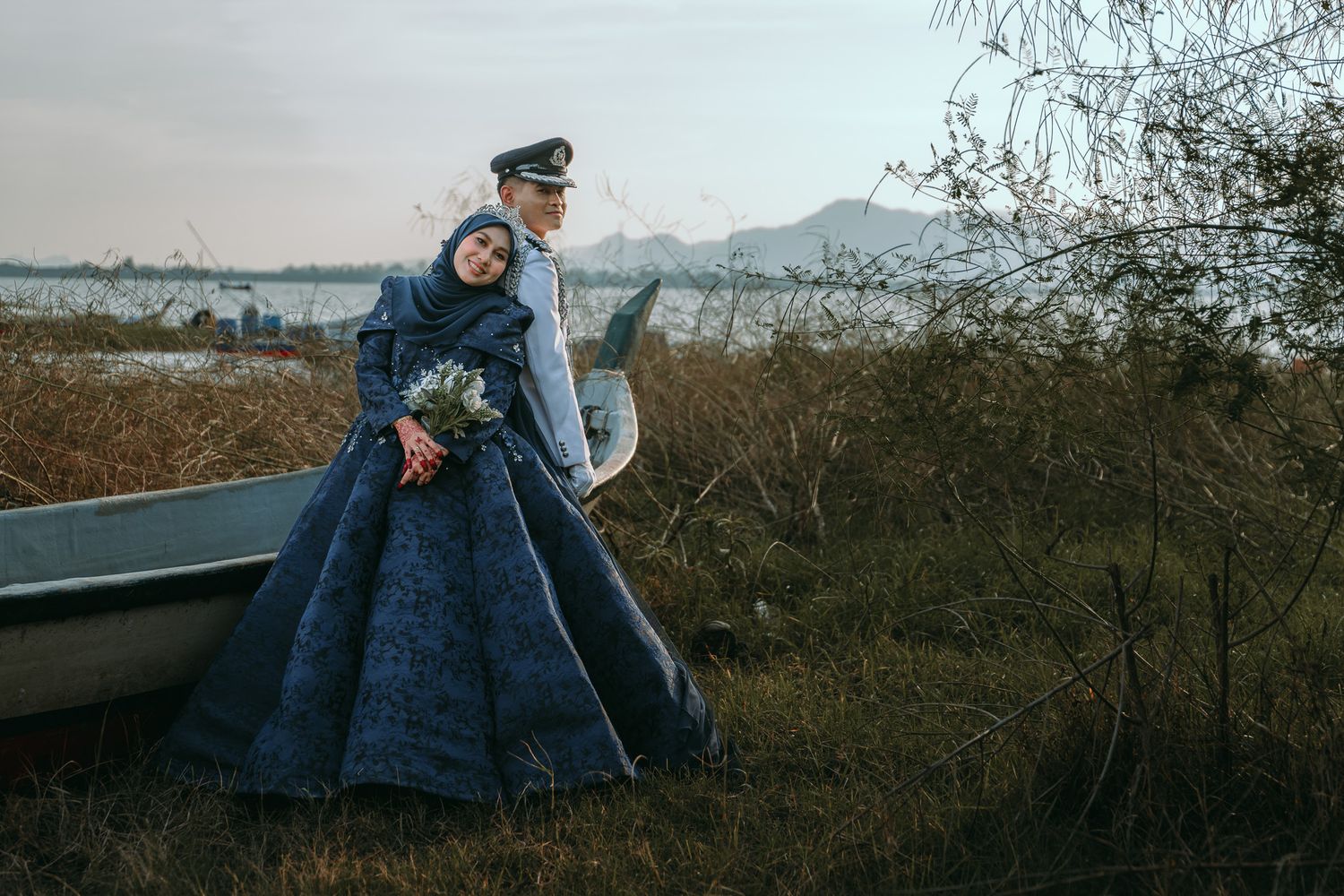 A couple in vintage winter coats embraces while standing in a field during sunset with mountains in the background.