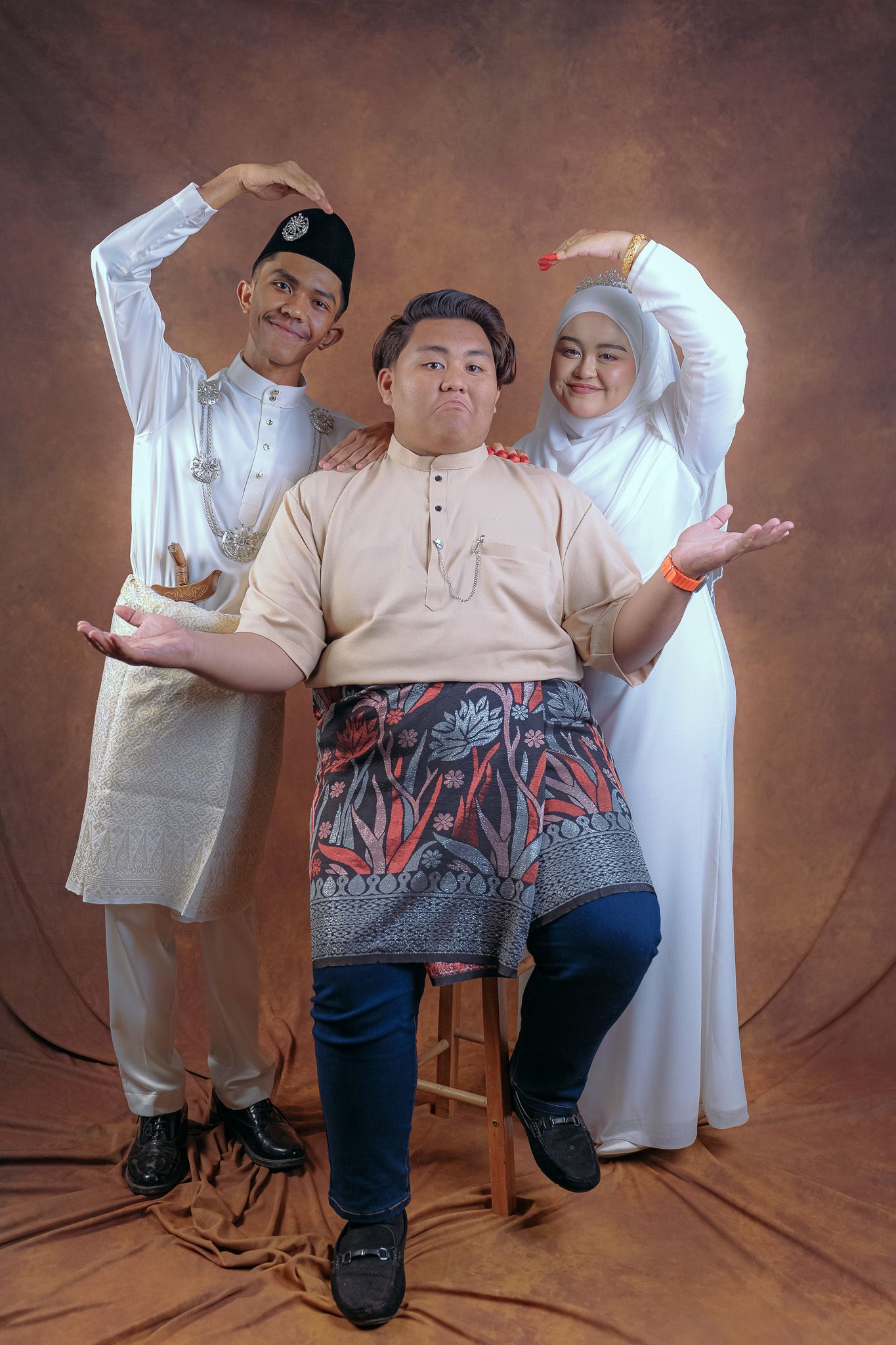 Three people in traditional Malay dress strike playful poses against a brown studio backdrop.