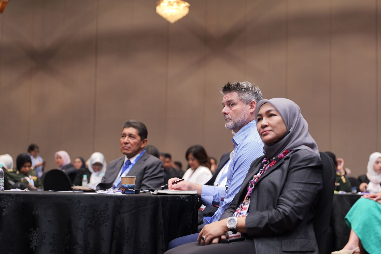 Conference participants seated at black-clothed tables listening intently to a presentation.