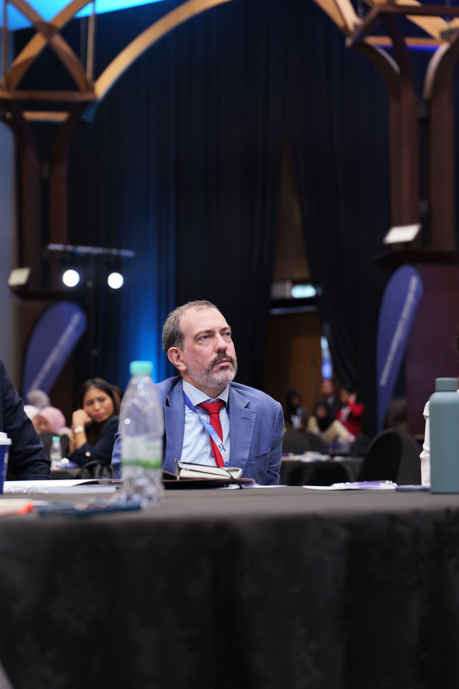 Professional conference attendee in a blue suit sits at a round table during a business event in an elegant venue.