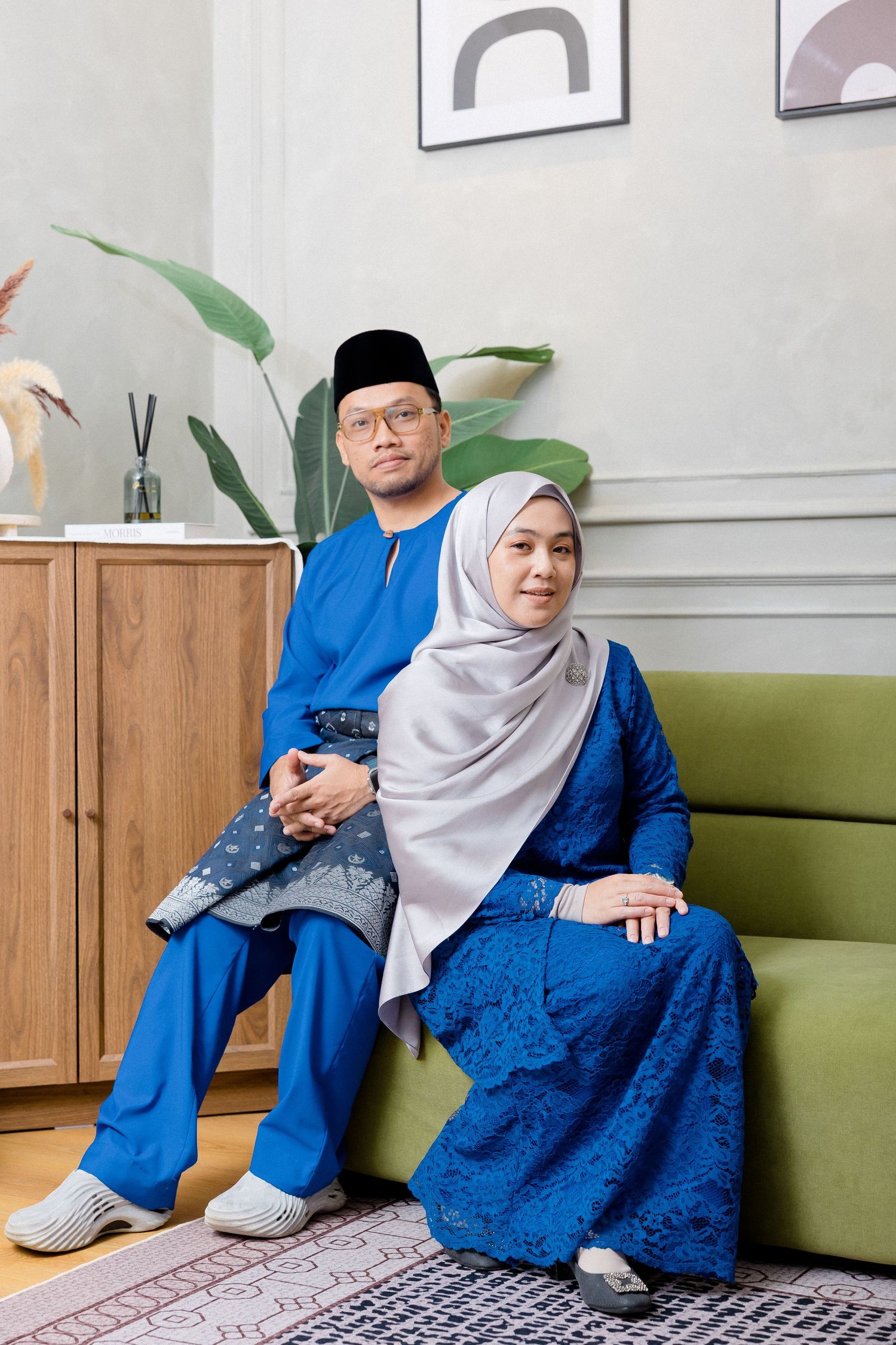 Person in matching royal blue outfit and light gray hijab sits on green couch in living room setting.