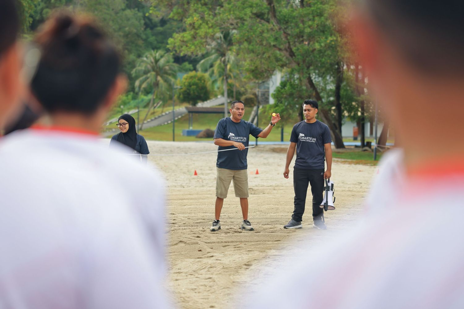 Coach instructs athletes on sandy beach training ground surrounded by players in white uniforms.