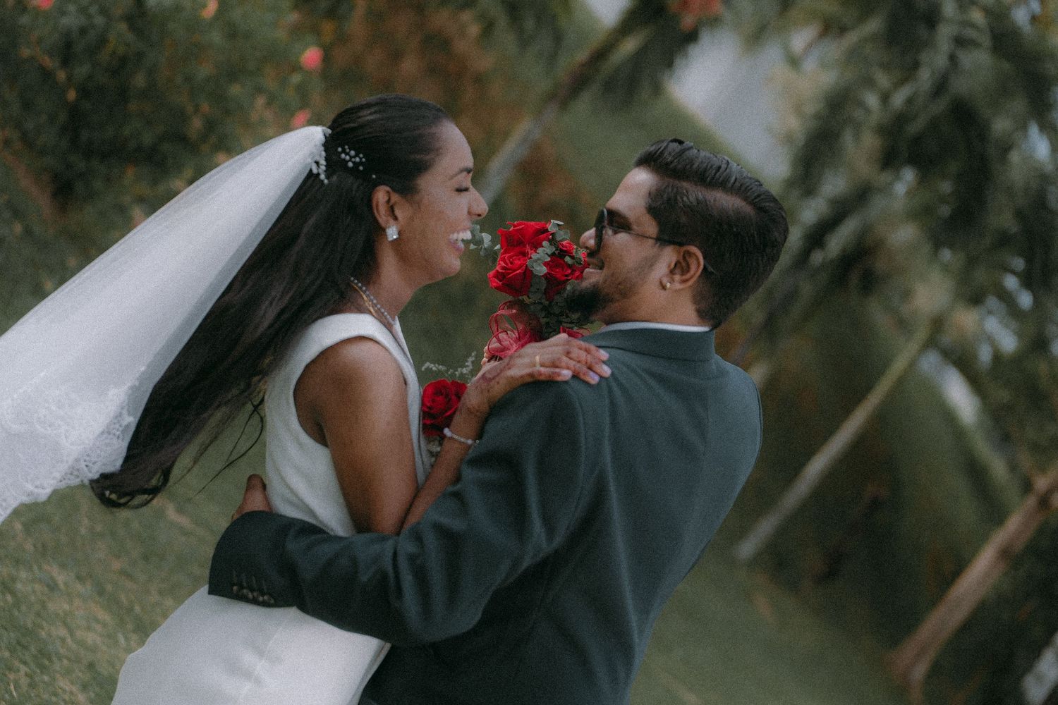 A newlywed couple shares a playful moment with red flowers in an outdoor garden setting.