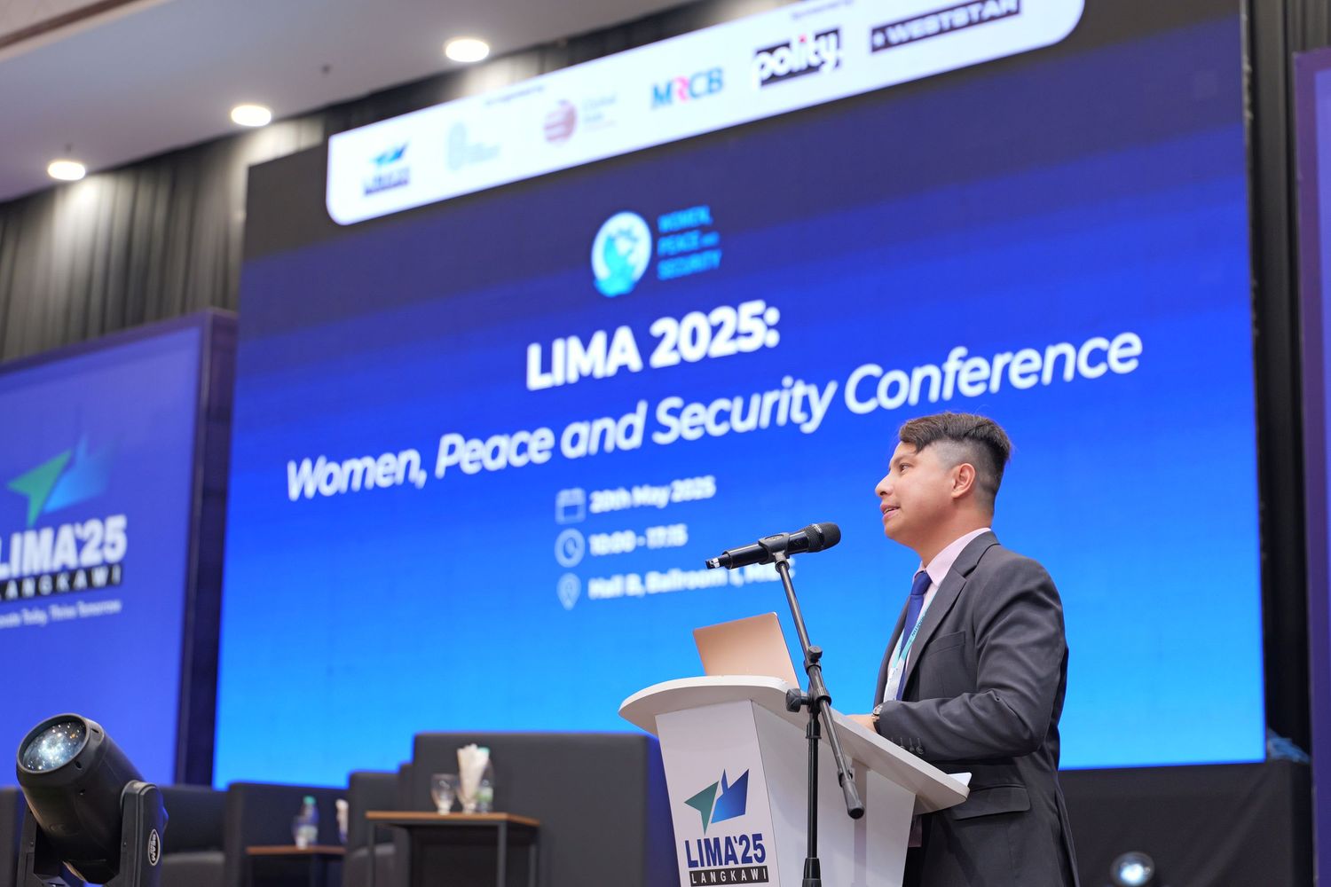 Large presentation screen displays event title at conference venue with blue lighting and stage setup.