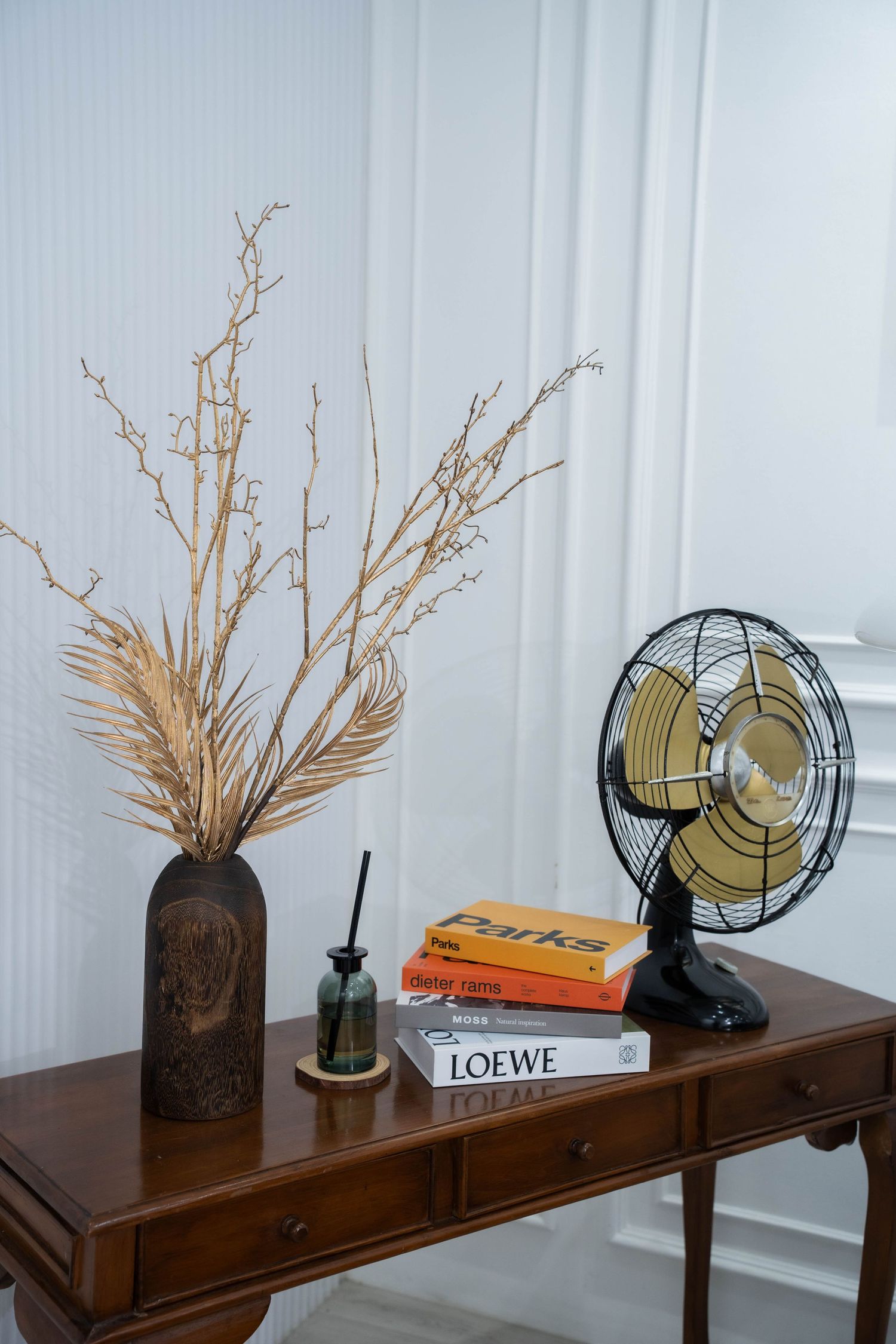 A vintage electric fan sits on a wooden console table alongside dried floral arrangements and decorative books.