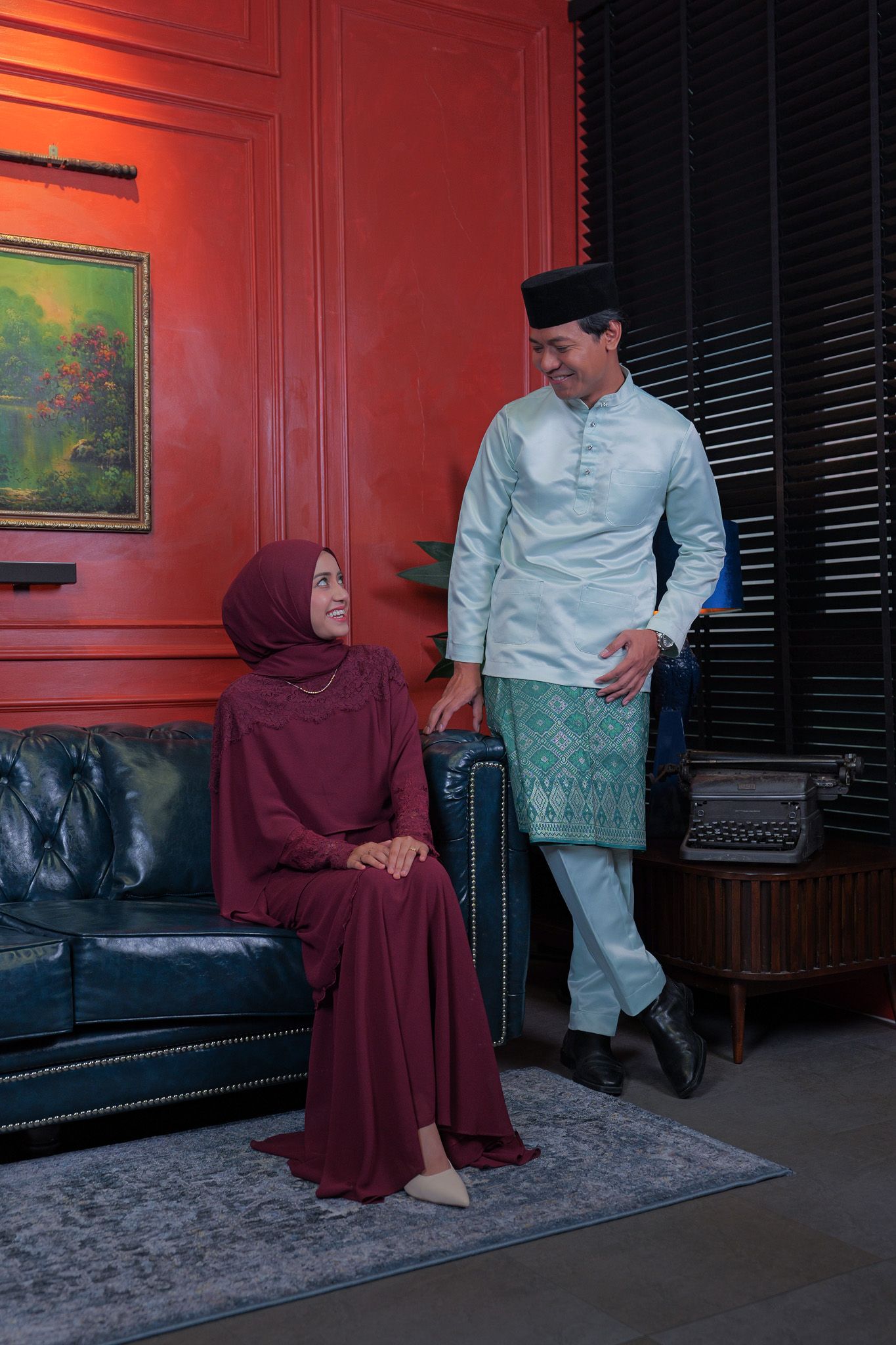 Two people sit in a room with red walls and dark furniture while having a conversation in traditional attire.