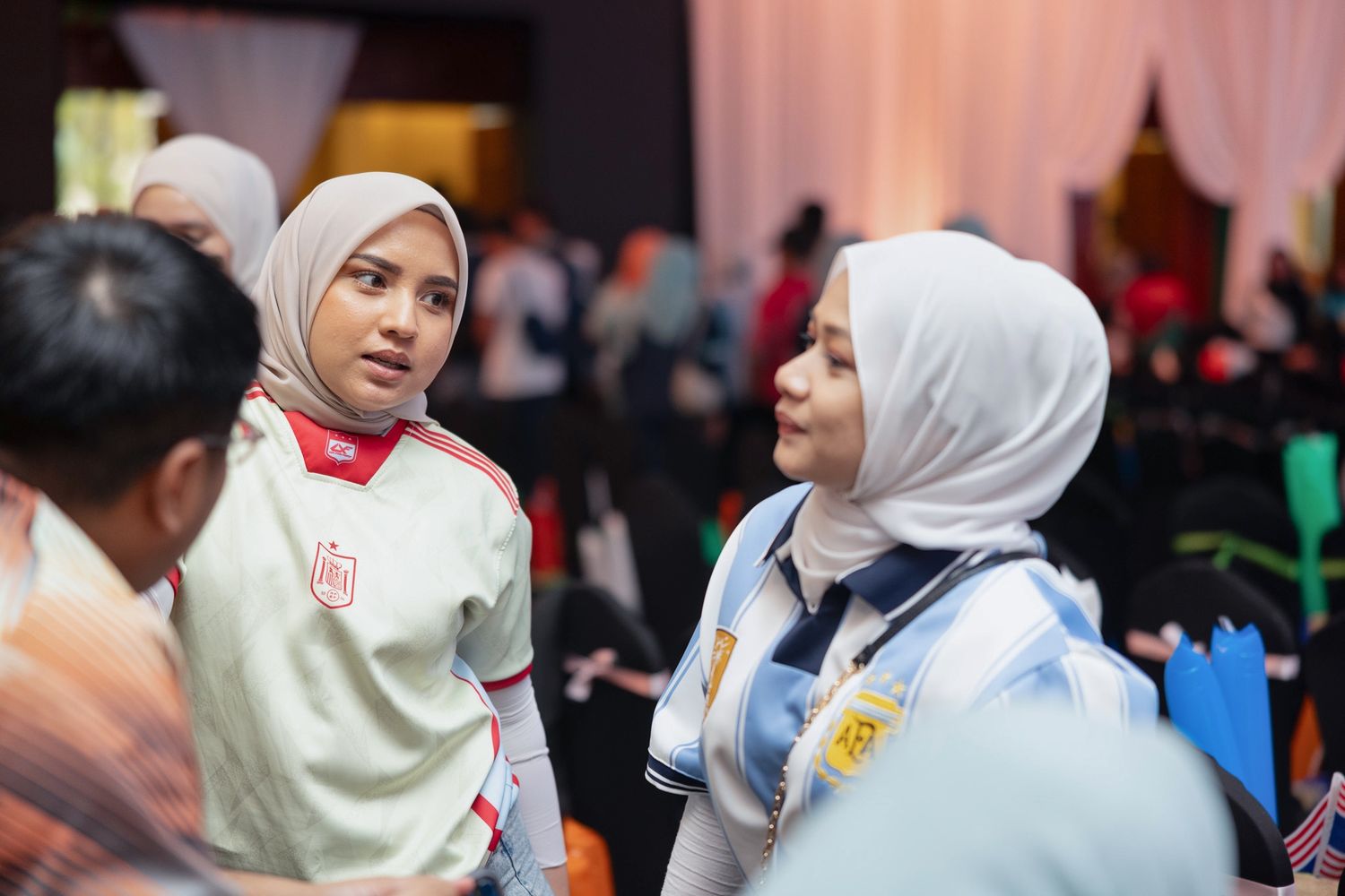 Two women wearing hijabs engage in conversation at an indoor event, one in a red uniform and one in a white uniform.
