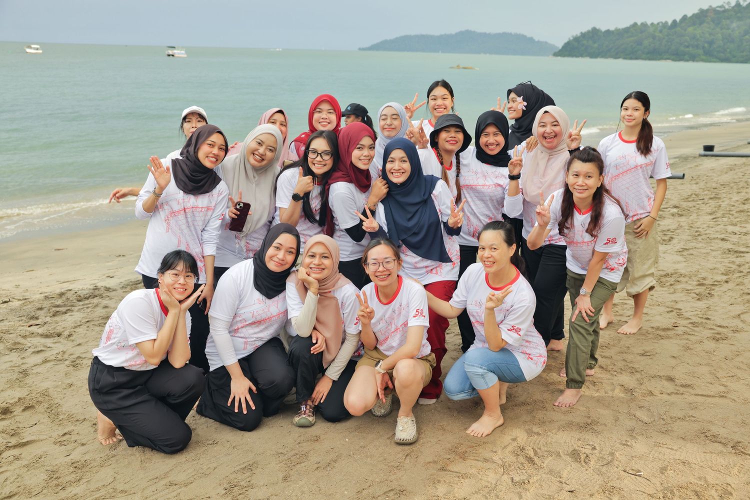 Group of women posing together on a tropical beach, wearing matching white shirts, smiling and making peace signs.