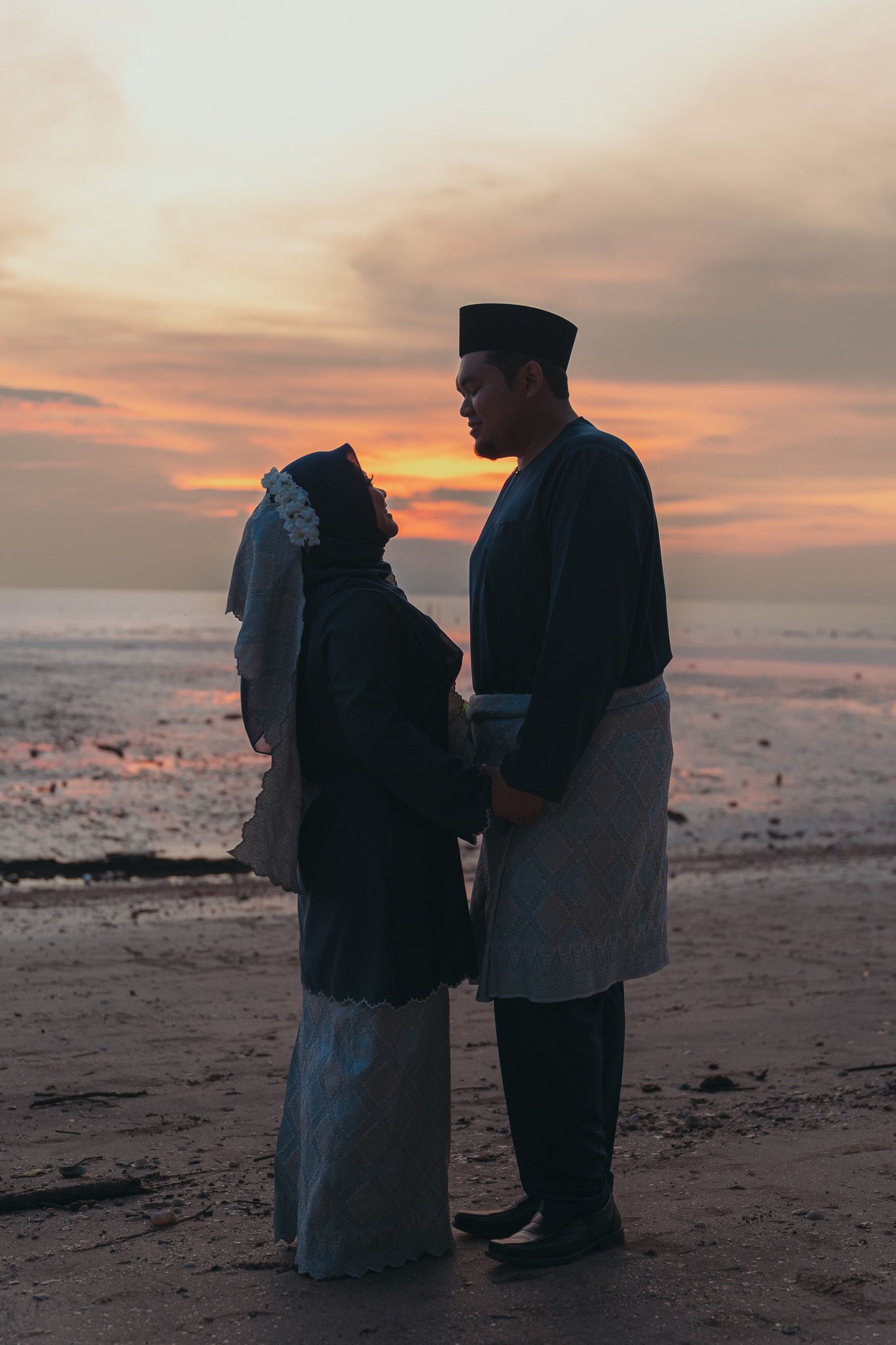 A couple in traditional Muslim attire stands silhouetted on a beach during a beautiful sunset over the ocean.