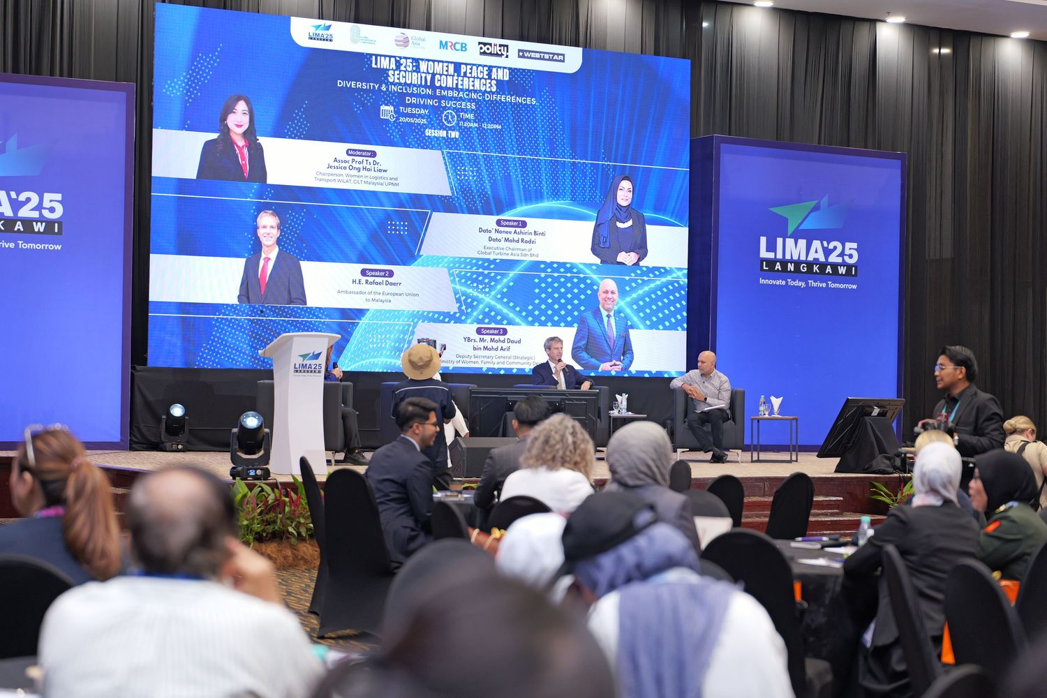 Large video screens display presentations at a LIMA conference while attendees watch from their seats.