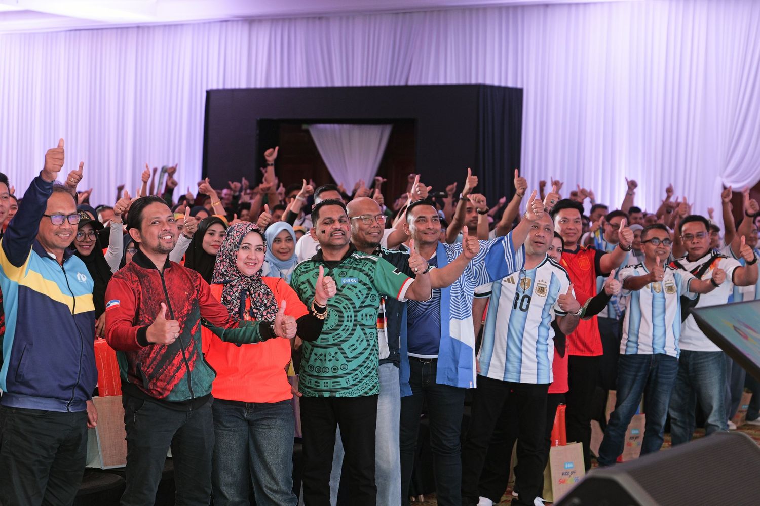 Diverse crowd raises hands and cheers together at an energetic indoor gathering or conference.