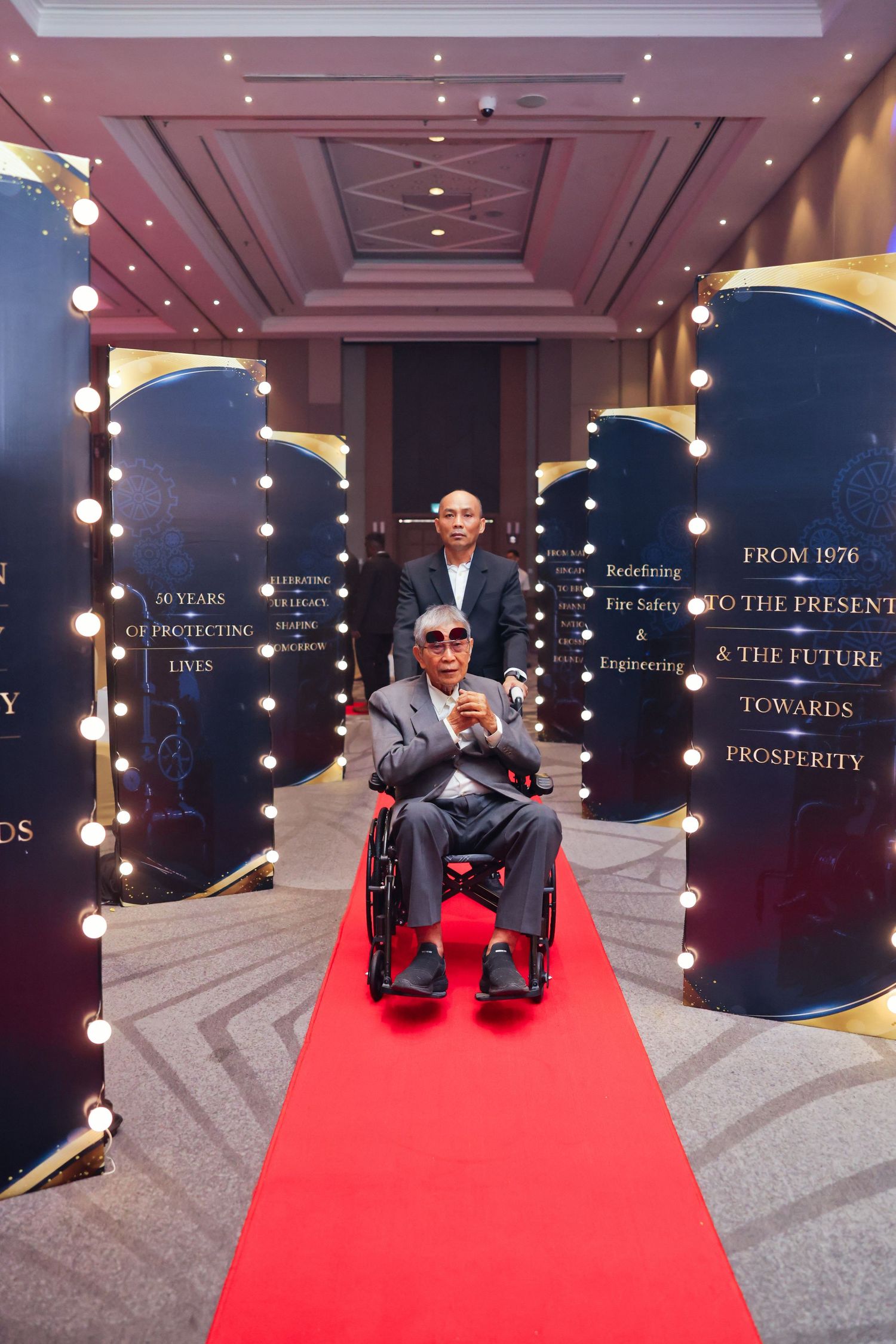 Elderly man in wheelchair on red carpet at formal event, escorted by attendant through illuminated display panels.