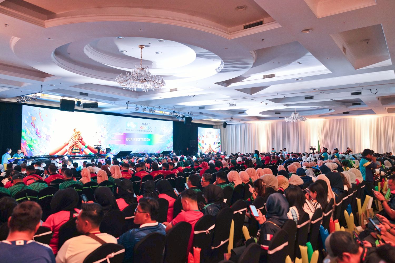 Large conference hall filled with attendees in colorful attire watching a vibrant presentation on a large stage screen.
