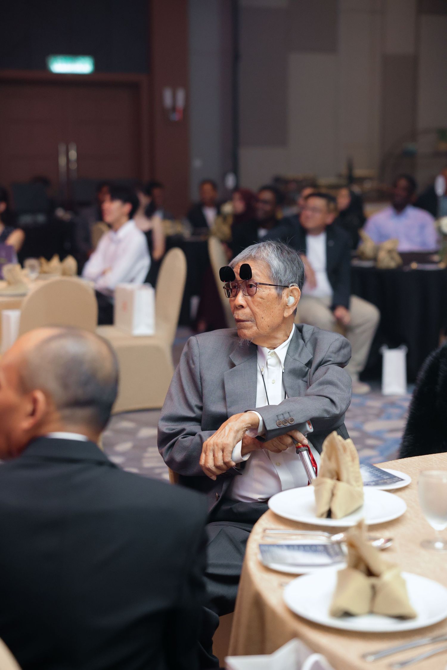 An elderly man wearing large sunglasses sits at a banquet table during a formal corporate event.