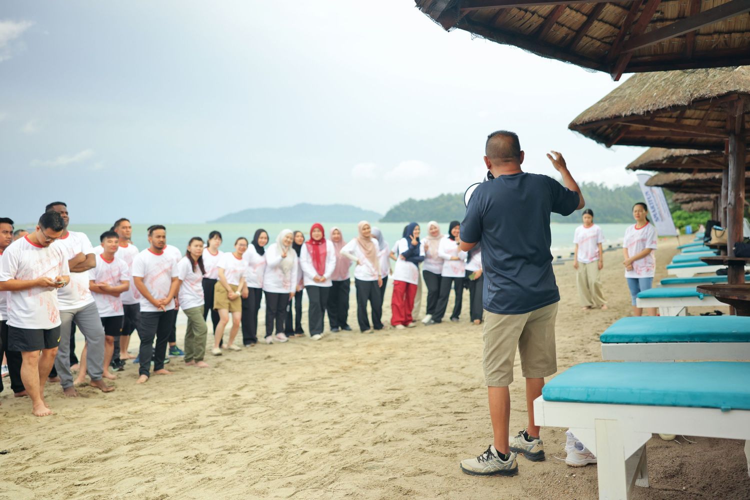 A group of people in white shirts listening to a speaker on a tropical beach with thatched umbrellas and turquoise water.