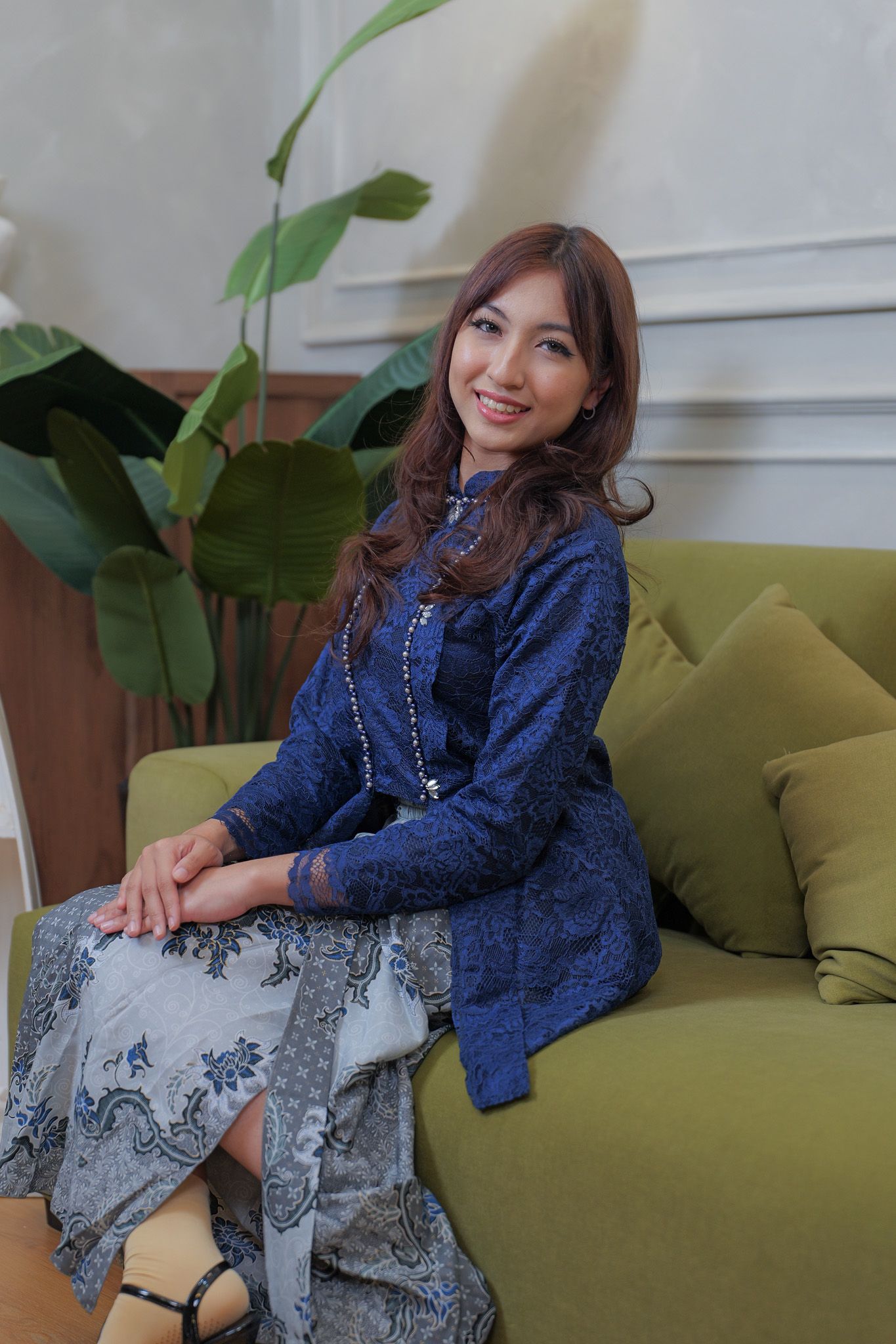 A person in a navy lace jacket and patterned skirt poses on a green couch with tropical plants in the background.