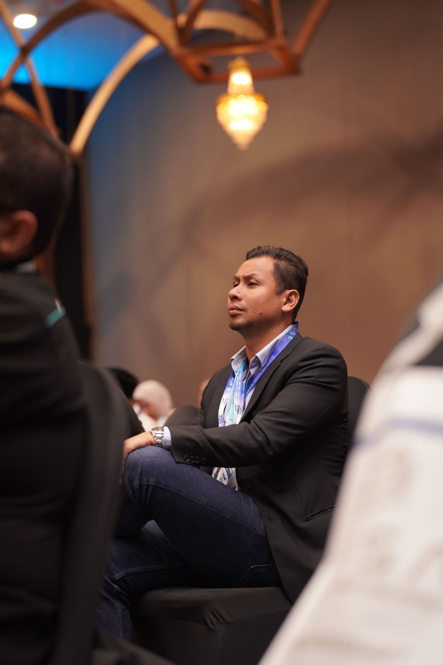 Conference attendee in business attire sits attentively during a presentation under decorative lighting.