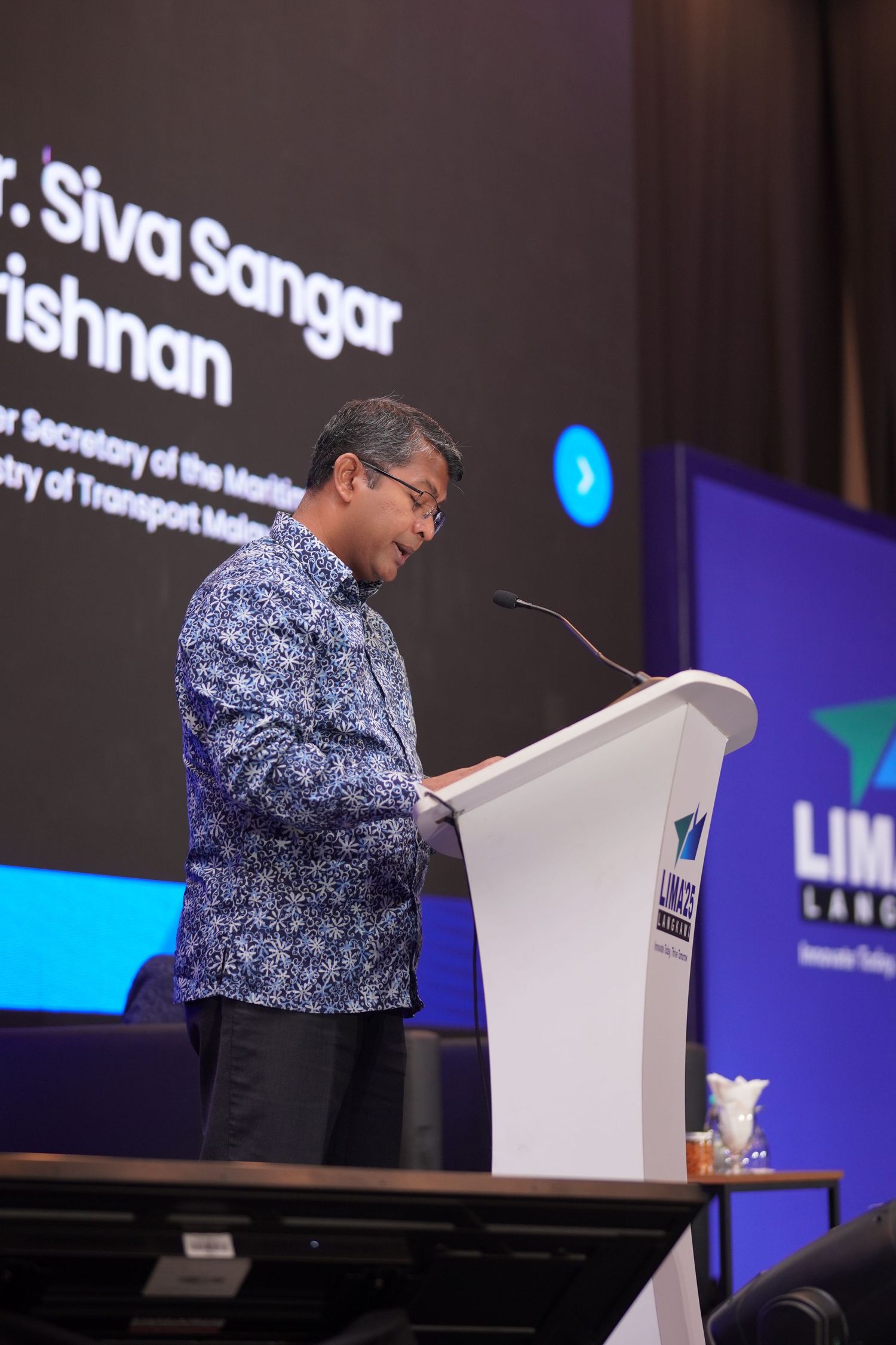 Speaker in traditional batik pattern shirt delivering presentation at podium during professional conference.