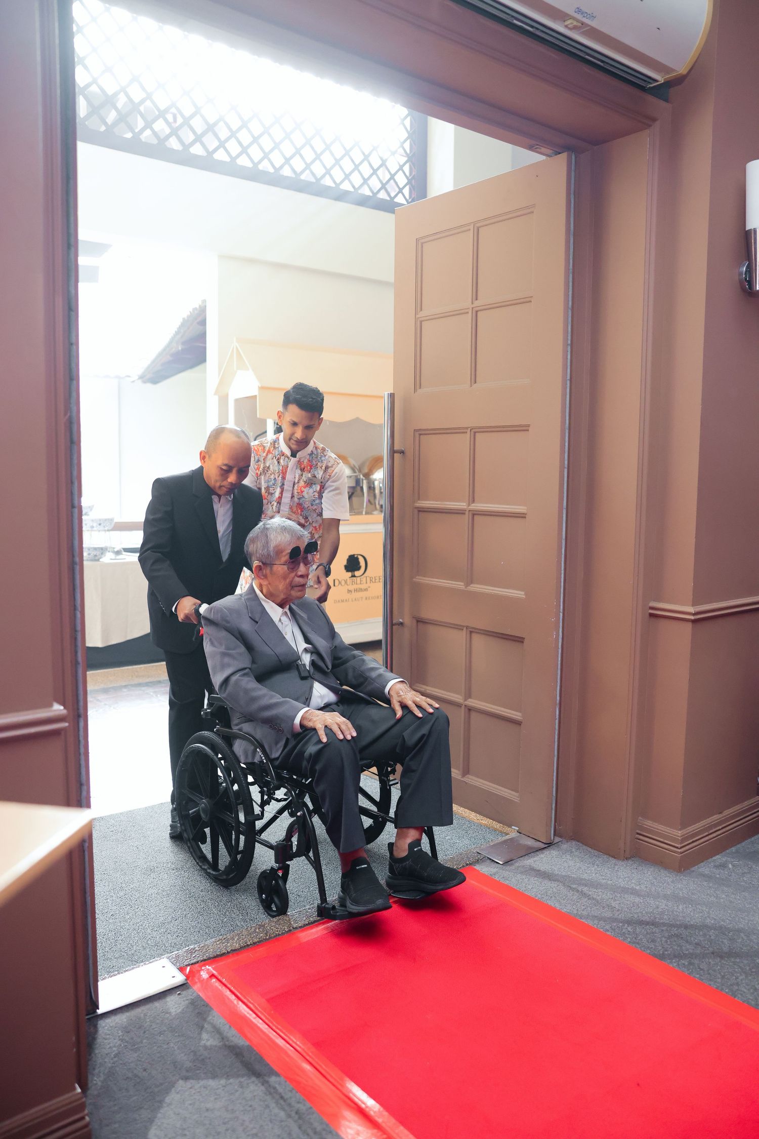 Elderly man in wheelchair being assisted through doorway with red carpet ramp by two attendants.