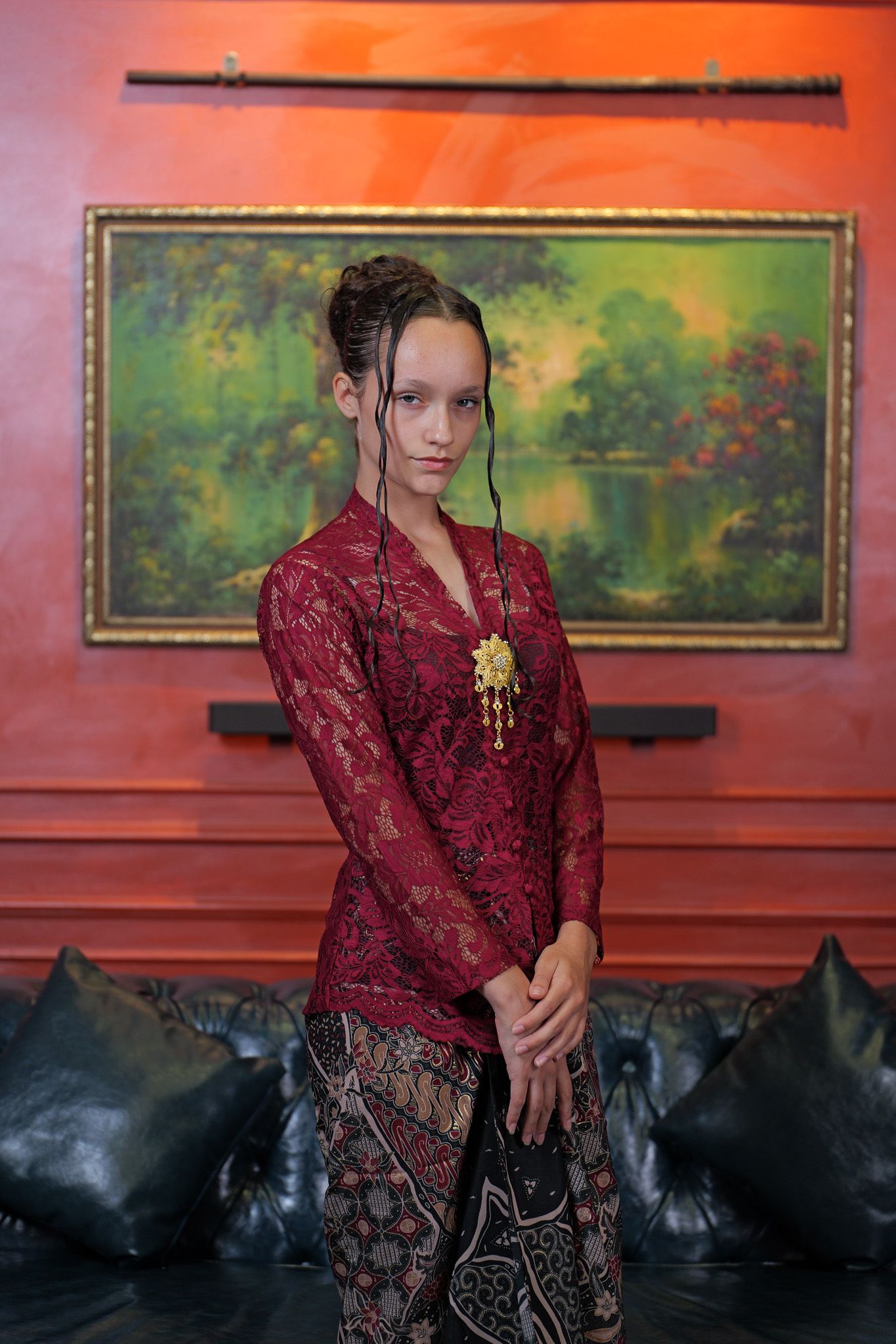 A figure in a burgundy lace top and patterned skirt poses against a red wall with artwork in the background.