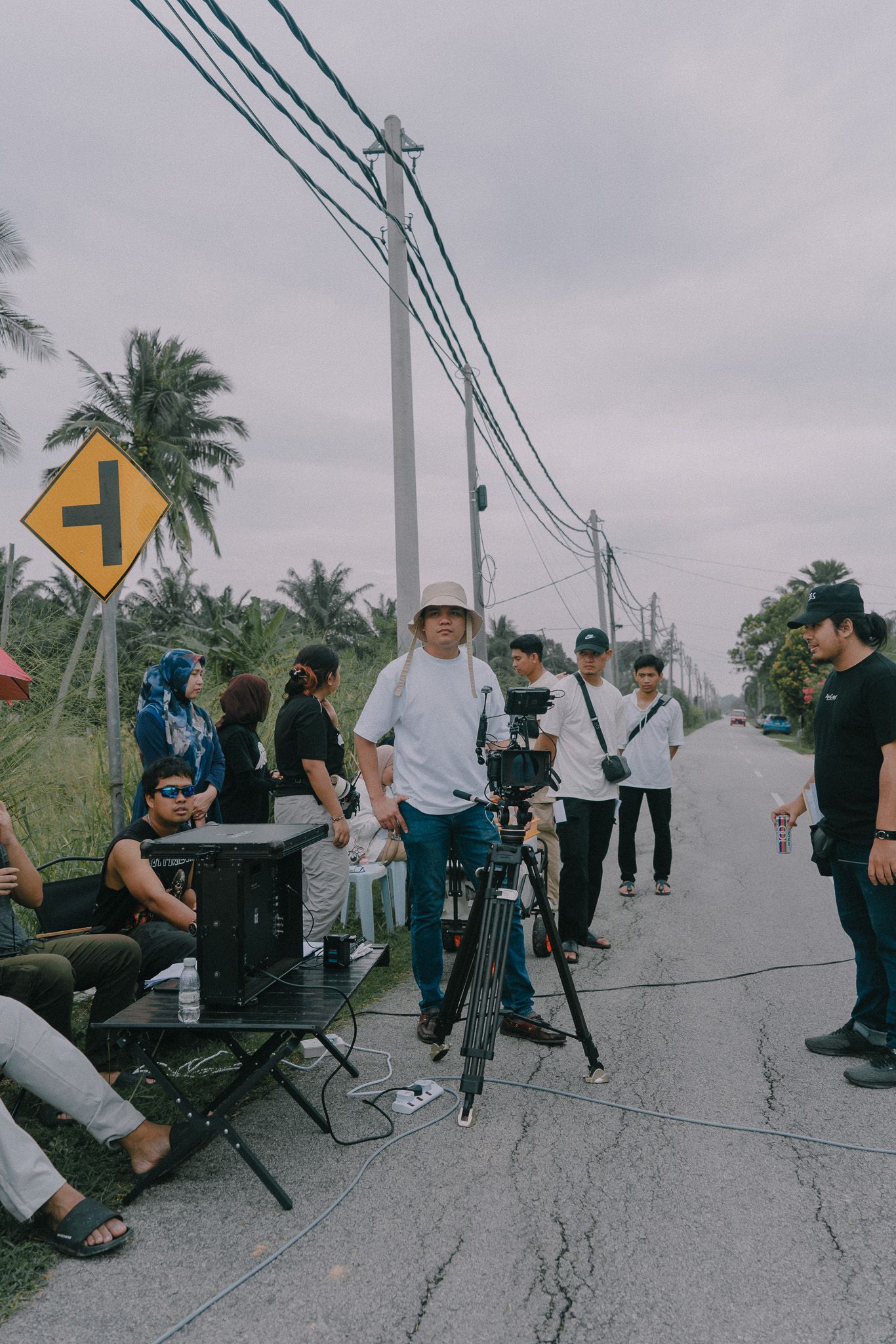 Film crew members stand with camera equipment on a rural road with palm trees and street signs in the background.