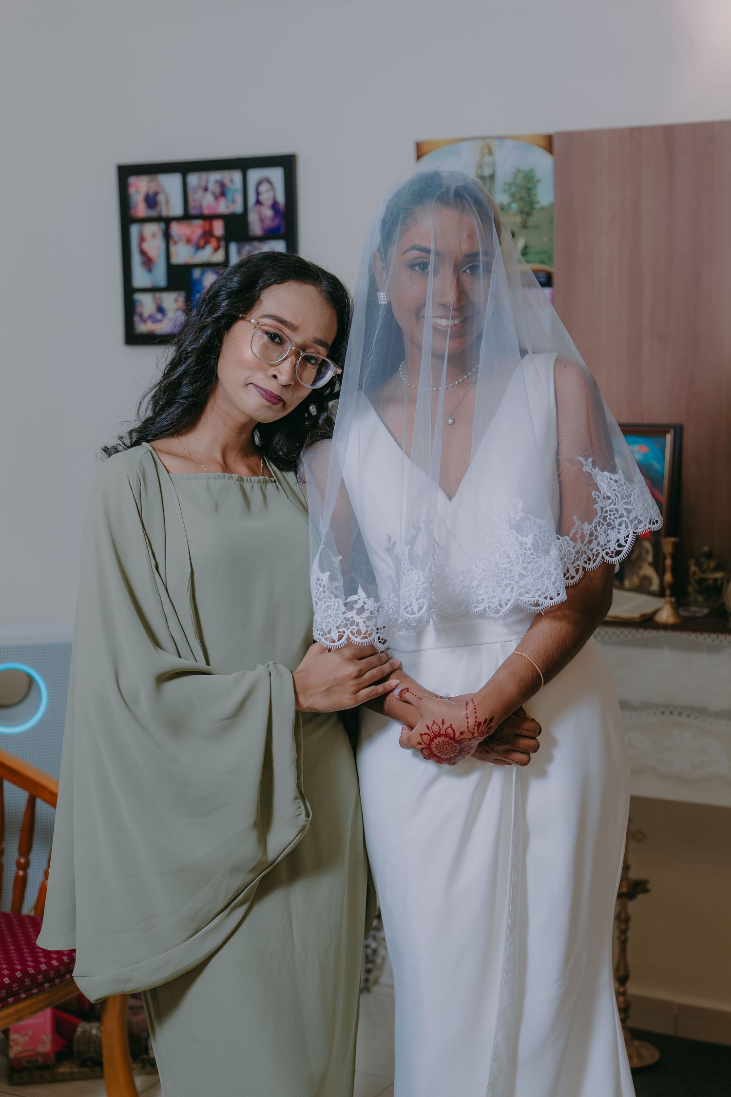 A bride in a white wedding dress and veil stands with someone wearing a sage green dress in a room with photos on the wall.