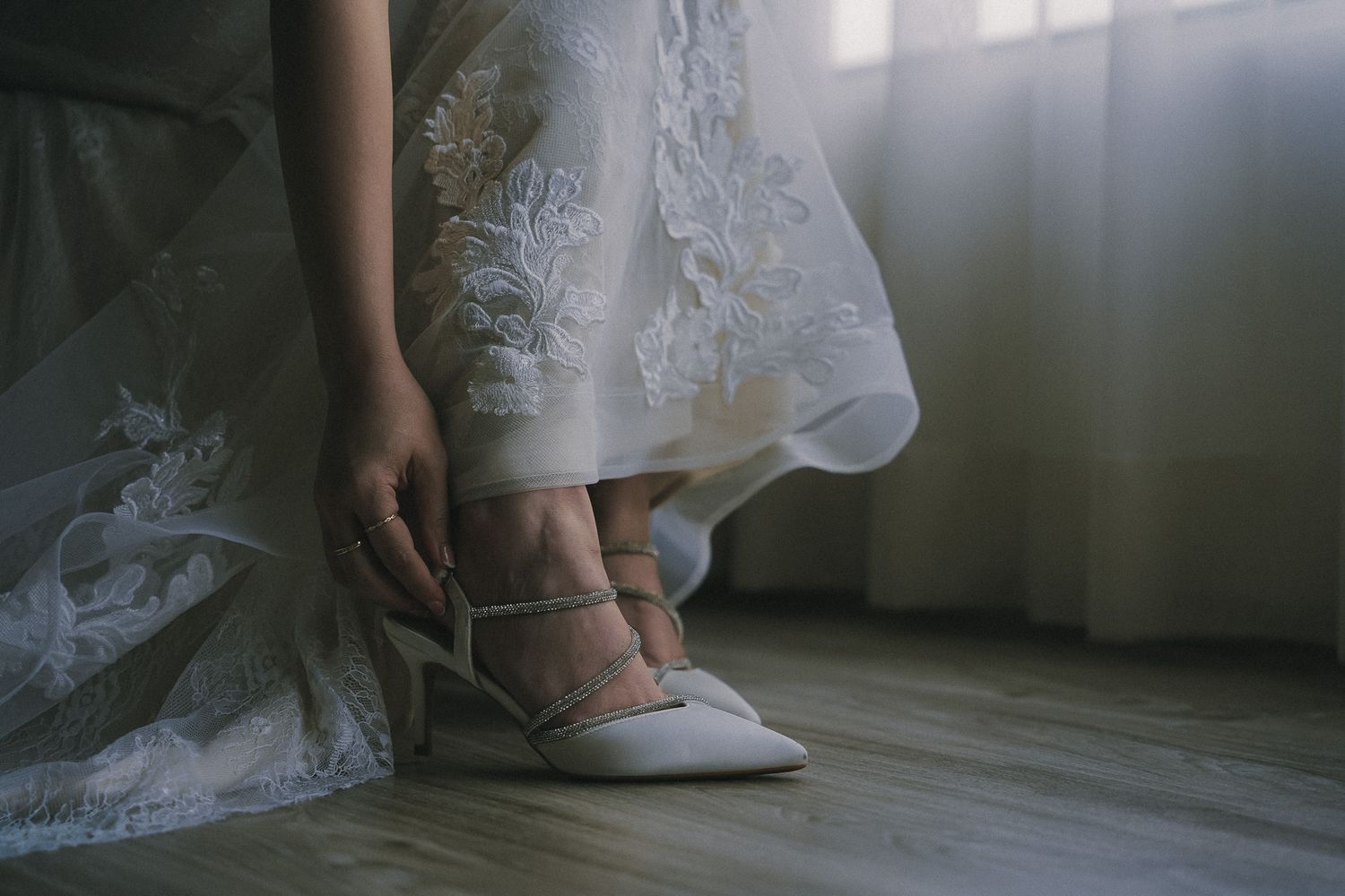 A bride adjusts her elegant white wedding shoes while wearing a lace wedding dress in soft natural light.