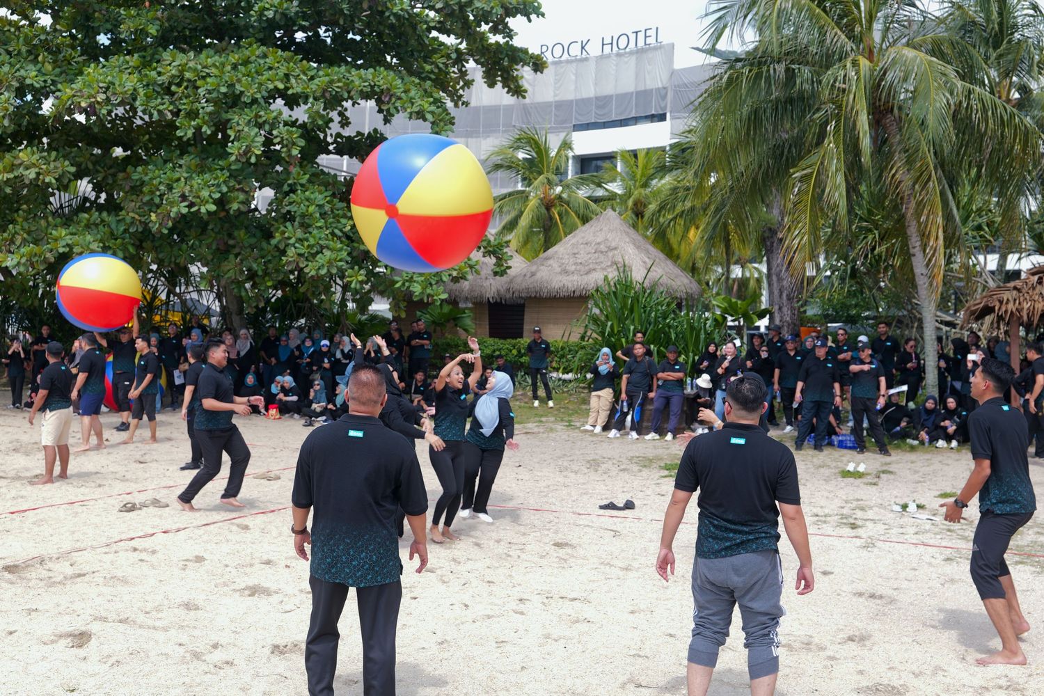 Large group playing with colorful beach balls on a sandy beach near a hotel during a team building event.